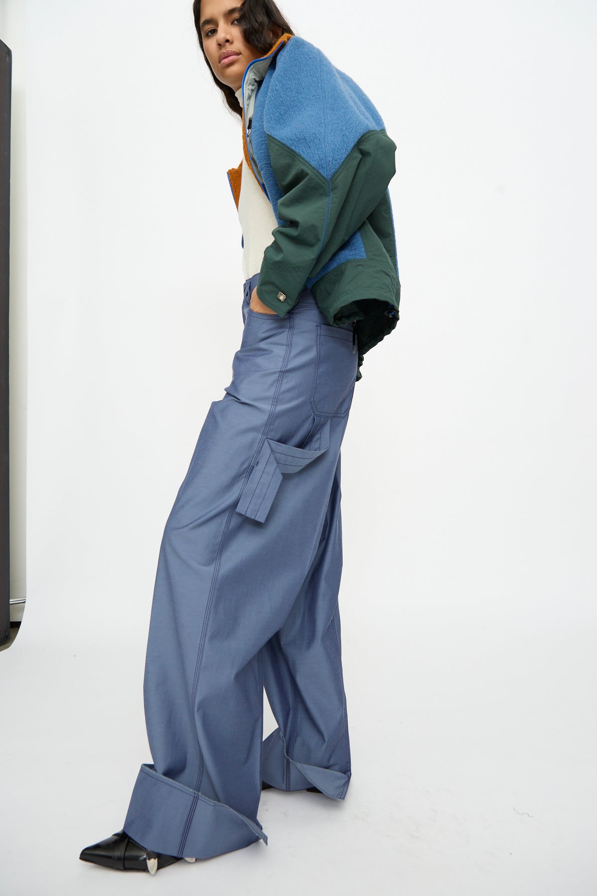 A person stands against a plain white background wearing an oversized color-block jacket, TOGA ARCHIVES Twill Painter Pants in Blue featuring a relaxed fit with large cuffs, and black pointed shoes.