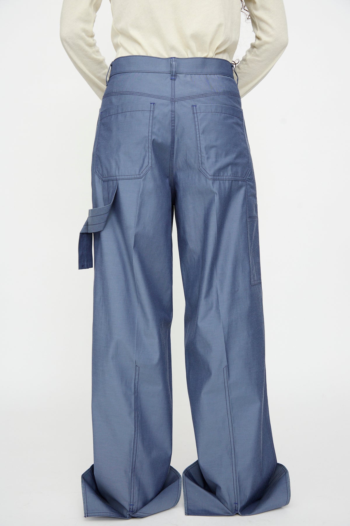 A person faces away, wearing a light-colored top paired with TOGA ARCHIVES Twill Painter Pants in Blue, featuring relaxed fit, wide cuffs, large back pockets, and a side strap detail, set against a plain background.