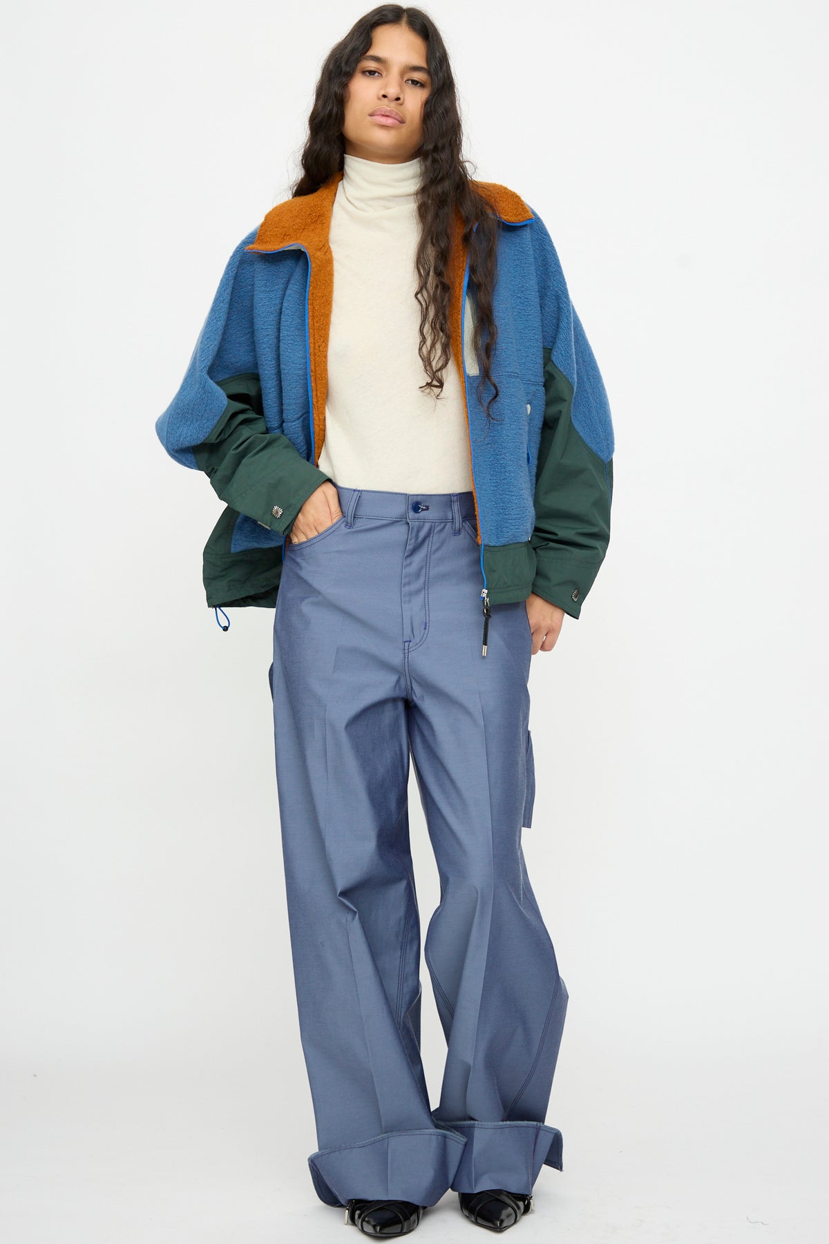A person stands against a plain background wearing a cream turtleneck, a relaxed fit blue and green color-block jacket with an orange collar, and the TOGA ARCHIVES Twill Painter Pants in Blue—wide-leg trousers featuring large cuffs inspired by classic painter pant details.