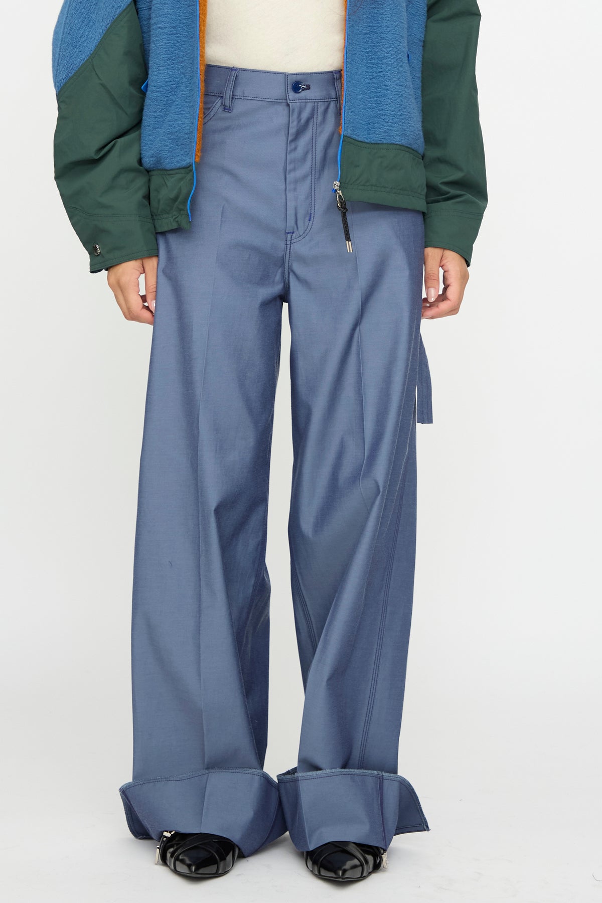 A person wearing TOGA ARCHIVES Twill Painter Pants in Blue, styled with black open-toe shoes, a white top, and a green and blue jacket, standing against a plain background.