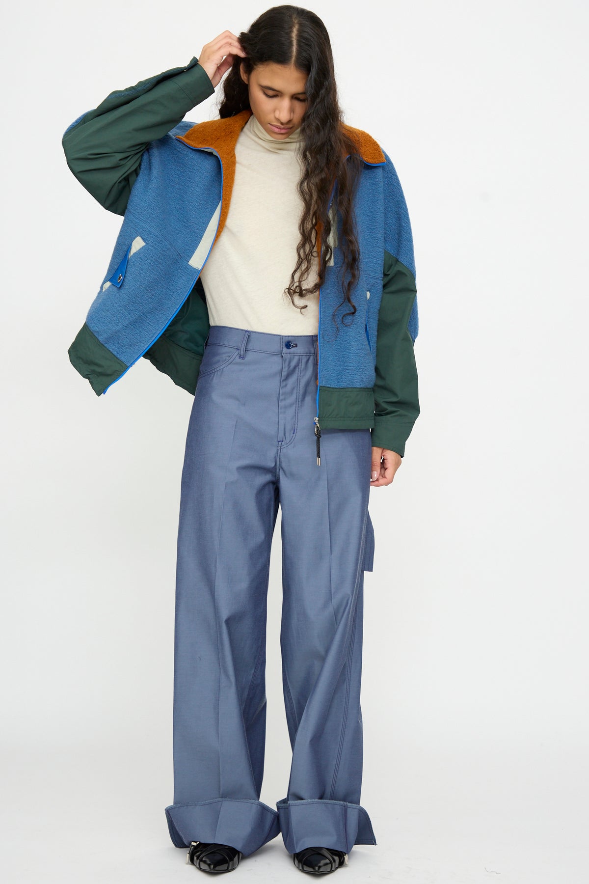 A person with long hair wears a cream turtleneck, a blue and green jacket, TOGA ARCHIVES Twill Painter Pants in Blue, and black shoes, standing against a plain white background.