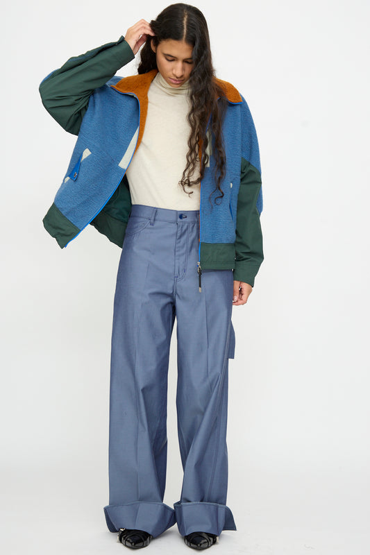 A person with long hair wears a cream turtleneck, a blue and green jacket, TOGA ARCHIVES Twill Painter Pants in Blue, and black shoes, standing against a plain white background.