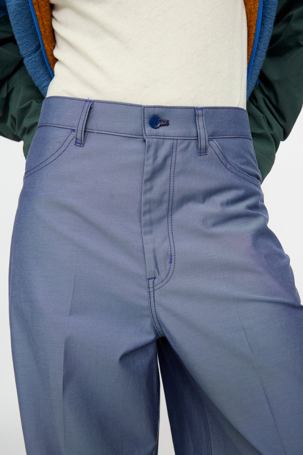 A person is shown from the waist down, wearing TOGA ARCHIVES Twill Painter Pants in Blue styled with a white shirt and a green jacket; focus is on the relaxed fit and high-waisted design of the pants, with the face not visible.