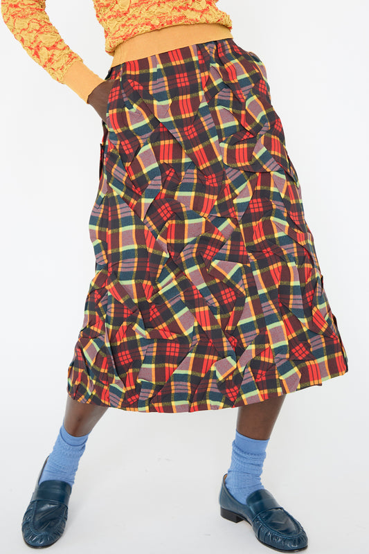 A person is wearing the Twill Plaid Skirt in Red from TOGA ARCHIVES, paired with a yellow and orange top, and blue socks and blue loafers.