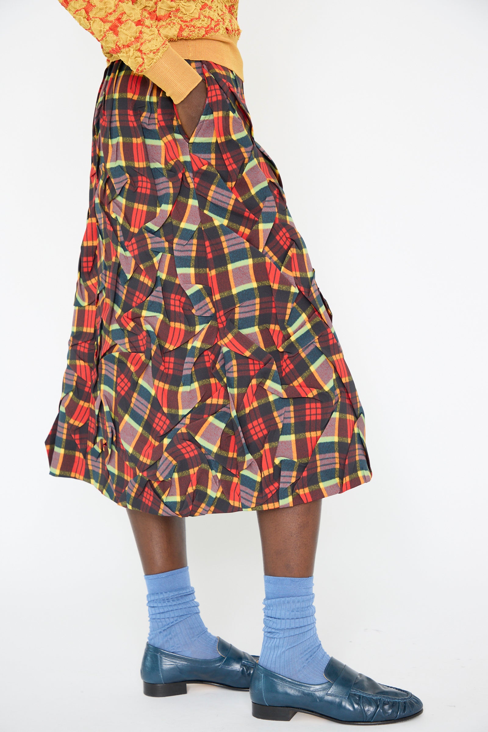 A person dons a high-waist, red plaid Twill Plaid Skirt by TOGA ARCHIVES, paired with light blue socks, blue loafers, and a yellow top.