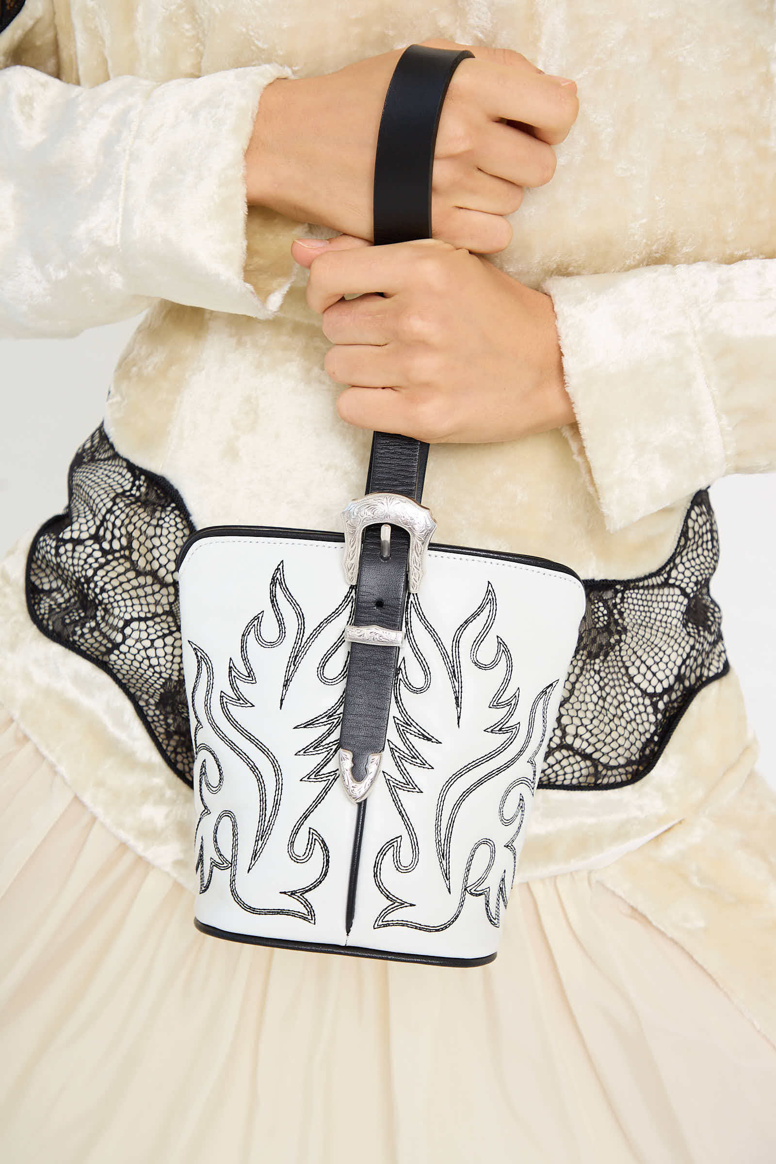 A person is holding the Western Leather Handbag in White by TOGA ARCHIVES, showcasing its black decorative patterns, black handle, and silver buckle detail. The purse is skillfully crafted from fine cowhide.