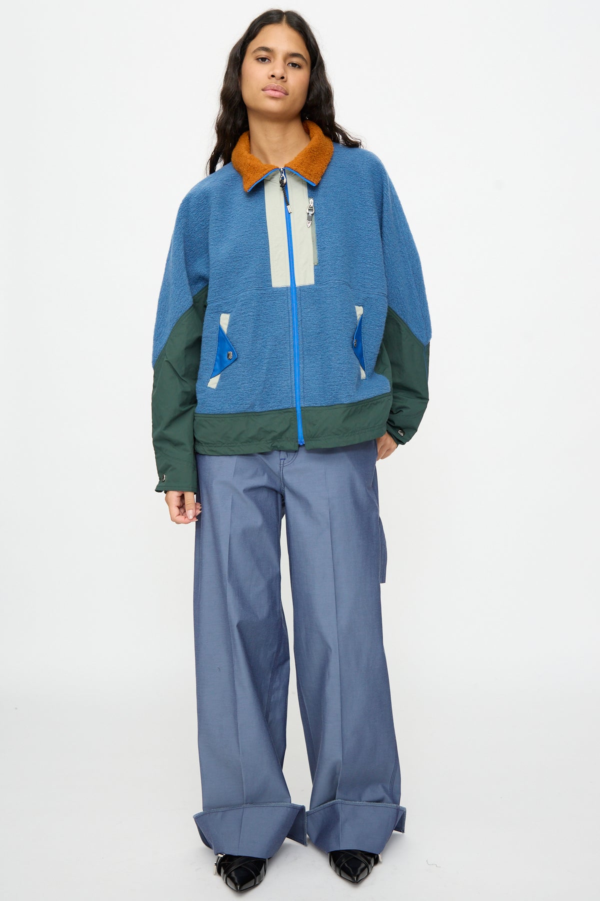 A person stands against a plain background wearing the TOGA ARCHIVES Wool Jersey Blouson in Light Blue—a relaxed fit jacket in blue and green with a brown collar, crafted from light blue wool and made in Japan—paired with wide-legged blue pants featuring folded cuffs and black shoes.
