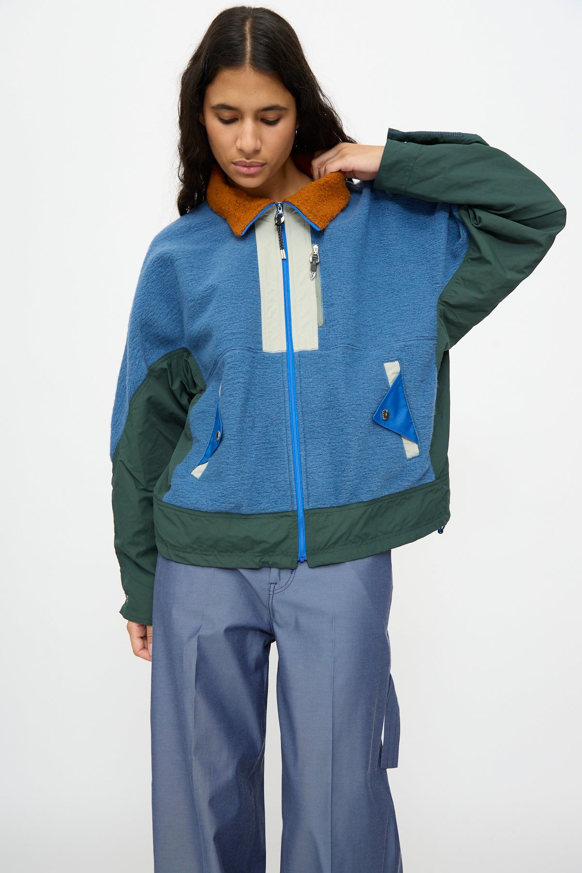 Wearing the TOGA ARCHIVES Wool Jersey Blouson in light blue—a relaxed fit jacket with blue and green tones, a brown collar, and crafted from Japanese wool—paired with wide-leg blue pants. The person stands against a plain background, looking down with one hand touching the back of their neck.