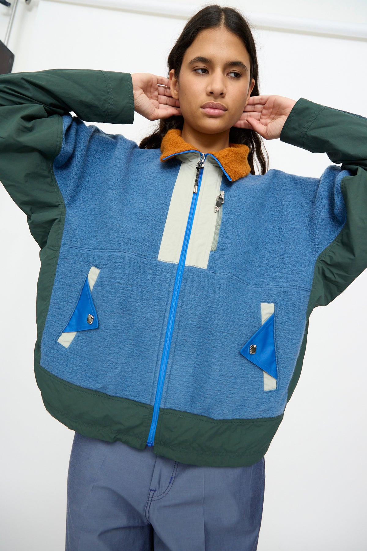 A person adjusts their collar while wearing the TOGA ARCHIVES Wool Jersey Blouson in Light Blue—a relaxed fit, color-block blue and green zip-up jacket featuring two front pockets, a brown collar accent, and crafted from light blue Japanese wool.