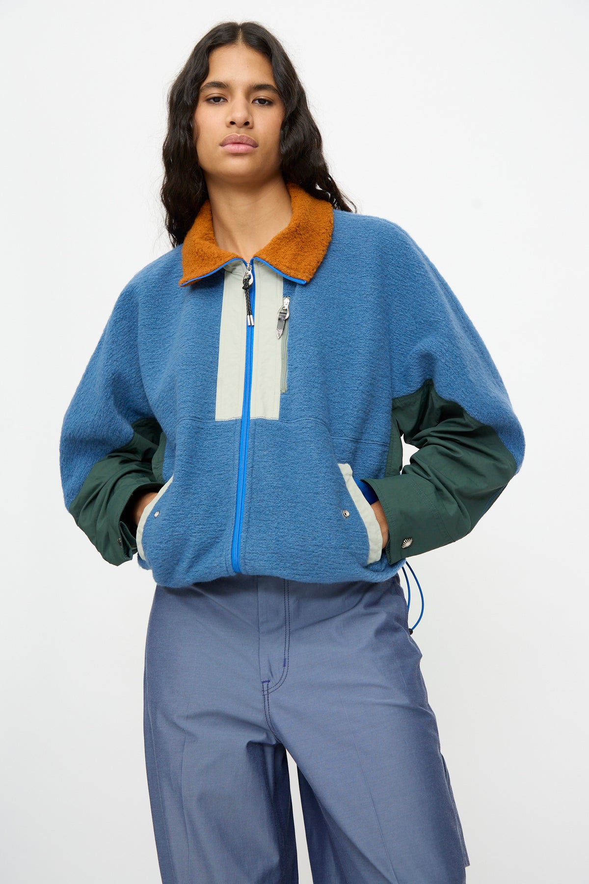 A person wearing the TOGA ARCHIVES Wool Jersey Blouson in Light Blue—featuring green sleeves, a tan collar, and a relaxed fit—paired with blue pants, made in Japan and photographed against a plain white background.