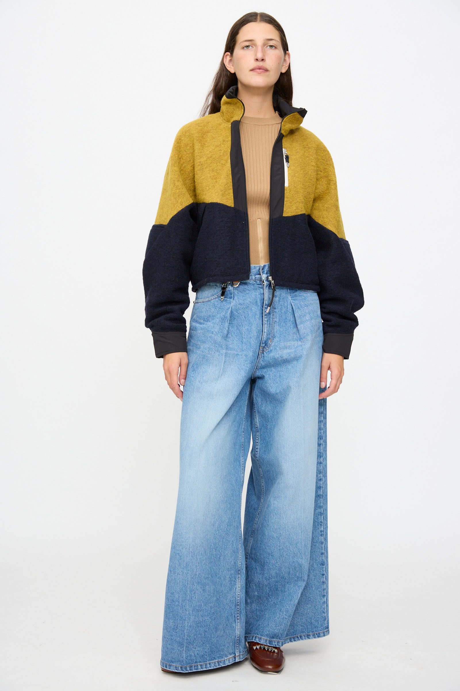 A person stands facing forward against a plain white background, wearing the TOGA ARCHIVES Wool Jersey Blouson in Navy styled with a tan ribbed top, wide-leg blue jeans, and brown shoes.