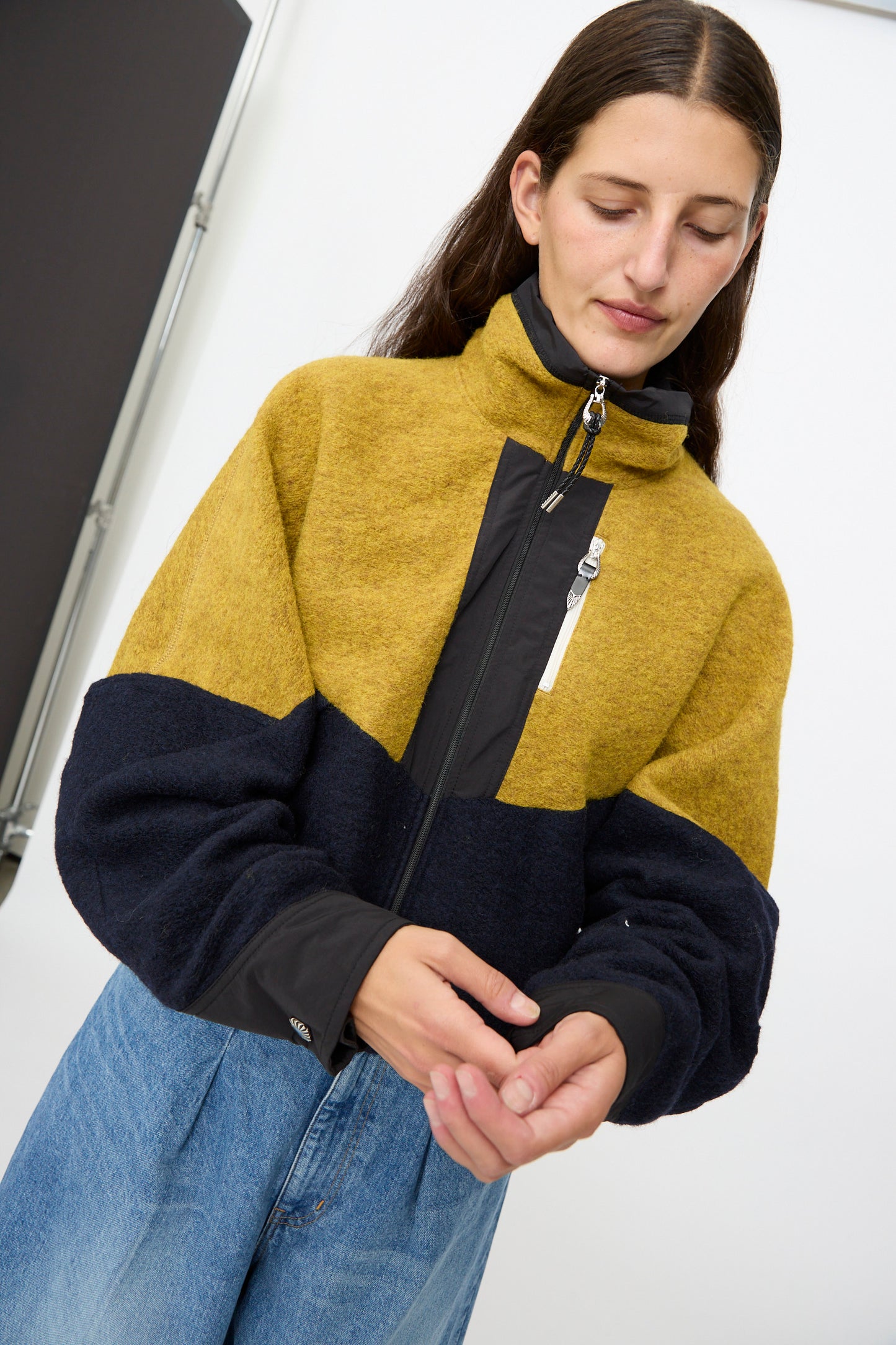 A person wearing the TOGA ARCHIVES Wool Jersey Blouson in Navy and blue jeans stands indoors with hands clasped, looking down.