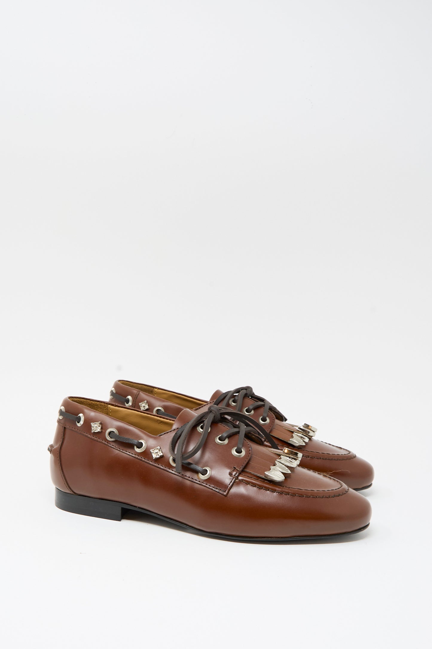 The TOGA ARCHIVES Leather Flat in Brown is a stylish pair of brown leather loafers featuring black laces, silver eyelets, metal embellishments, and classic fringe detail, shown against a plain white background.