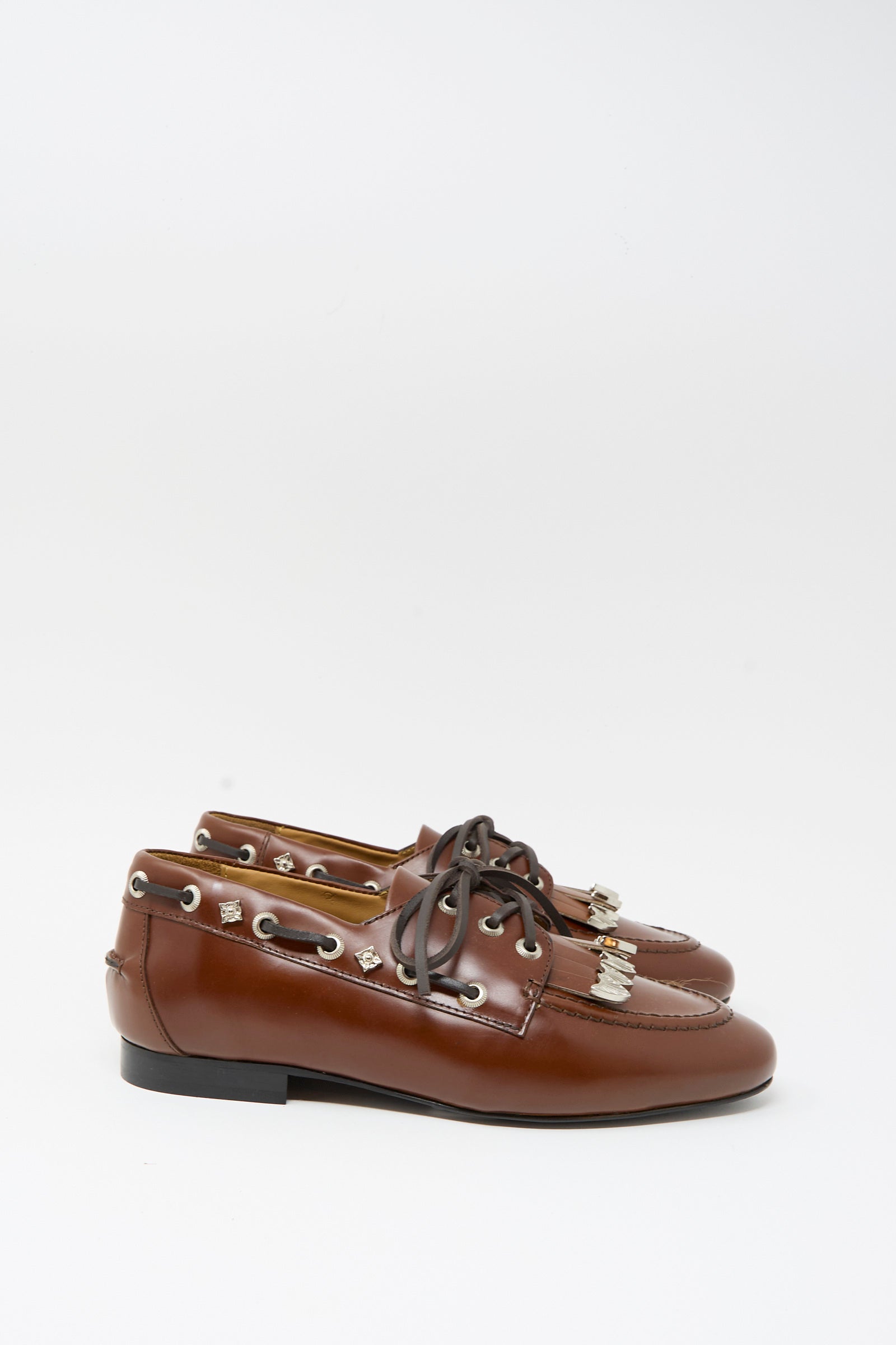 The TOGA ARCHIVES Leather Flat in Brown is a pair of brown leather boat shoes featuring black lace-up laces, silver eyelets, decorative metal accents, and subtle fringe detailing—displayed against a plain white background.