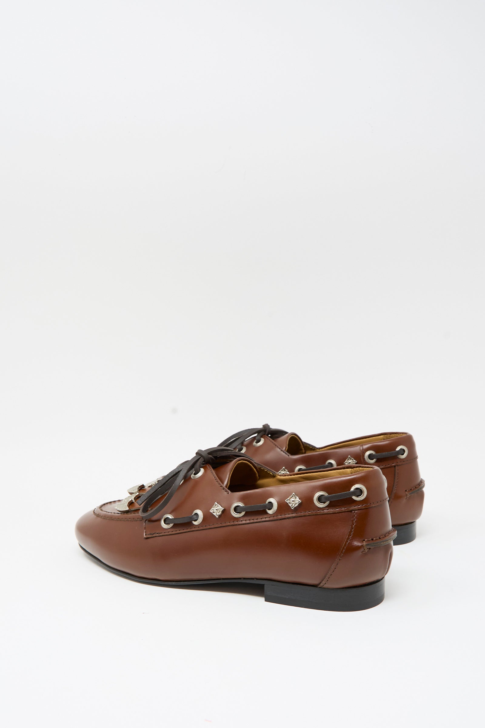 The TOGA ARCHIVES Leather Flat in Brown showcases refined brown leather with lace and tassel accents, silver eyelets, subtle fringe detailing along the top, and low black heels. Elegantly displayed against a plain white background, these classic flats embody timeless sophistication.