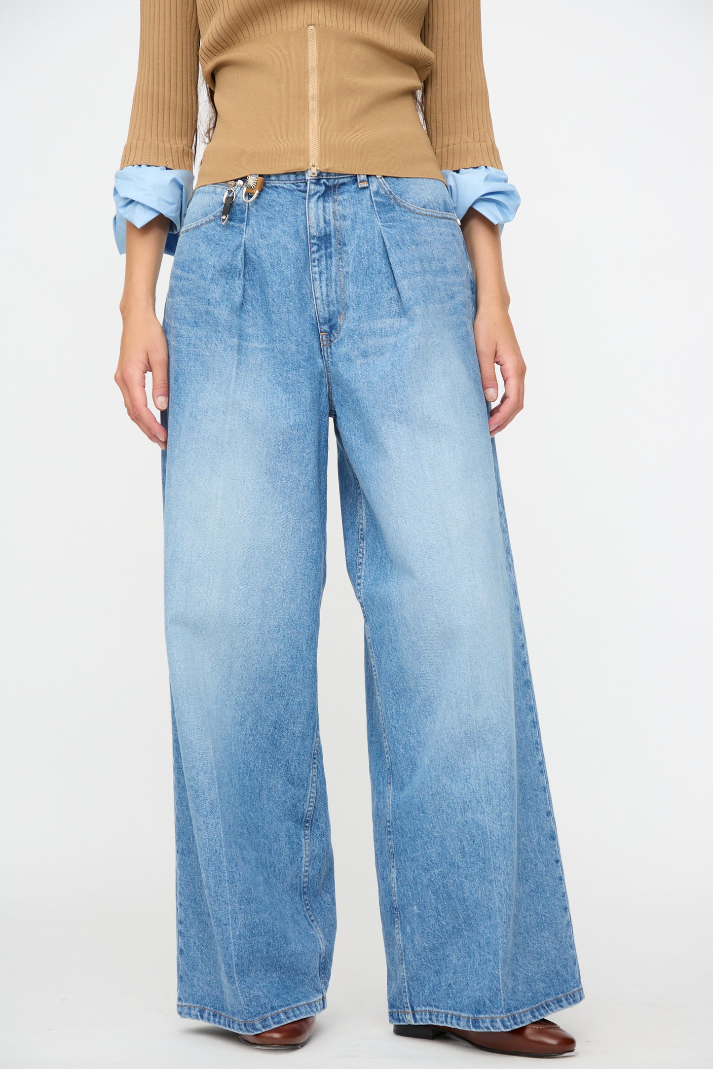 A person stands against a plain white background wearing TOGA ARCHIVES Denim Wide Pants in Blue—light blue, wide leg, mid-rise jeans made in Japan—paired with a tan ribbed top and a blue shirt with rolled-up sleeves.