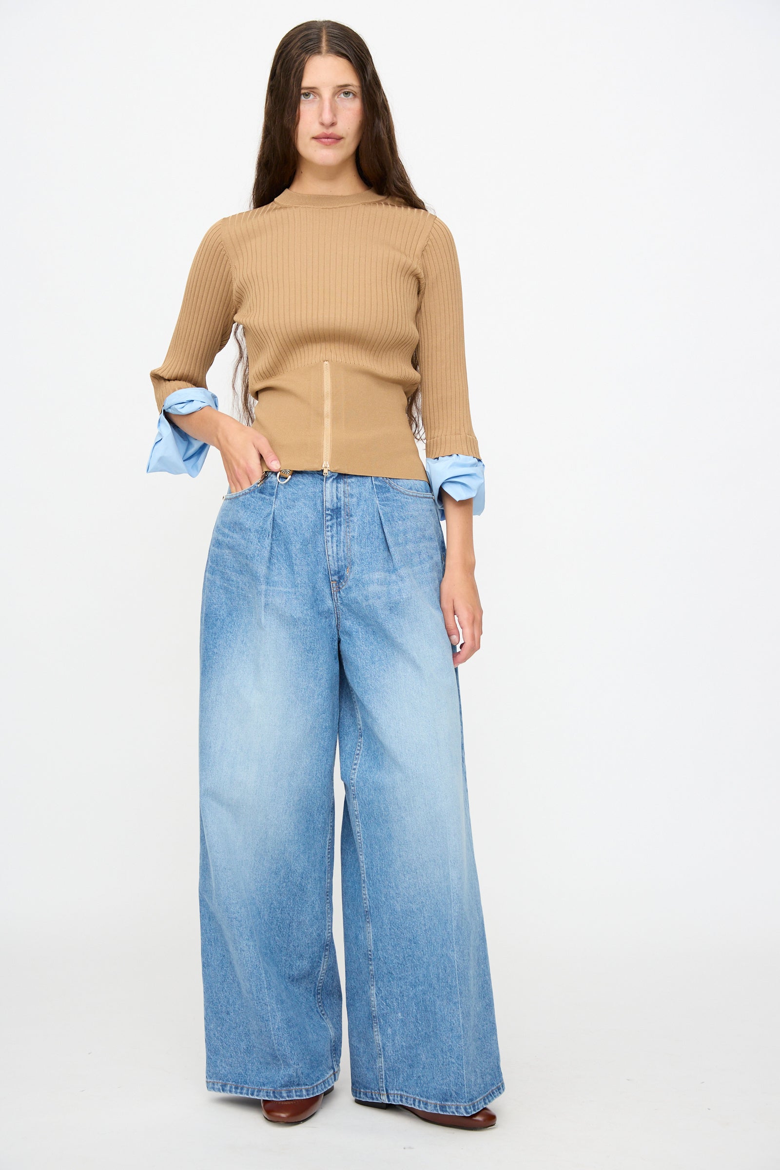 A woman stands against a plain background, wearing a ribbed tan long-sleeve top with blue cuffs, paired with TOGA ARCHIVES’ Denim Wide Pants in Blue—high-waisted, wide-leg jeans that bring a contemporary flair to her effortlessly chic ensemble. Brown shoes are partially visible.