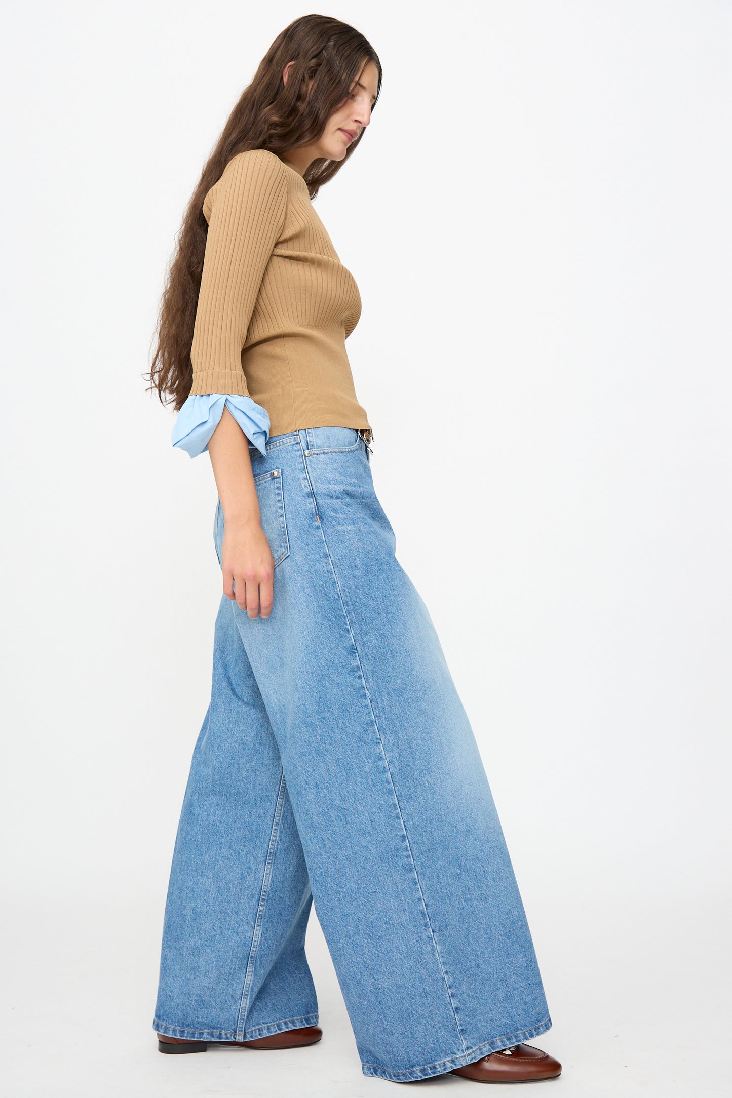 A person stands sideways against a plain white background, wearing a ribbed tan long-sleeve top with blue cuffs and the TOGA ARCHIVES Denim Wide Pants in Blue—high-waisted, wide-leg jeans crafted in Japan.