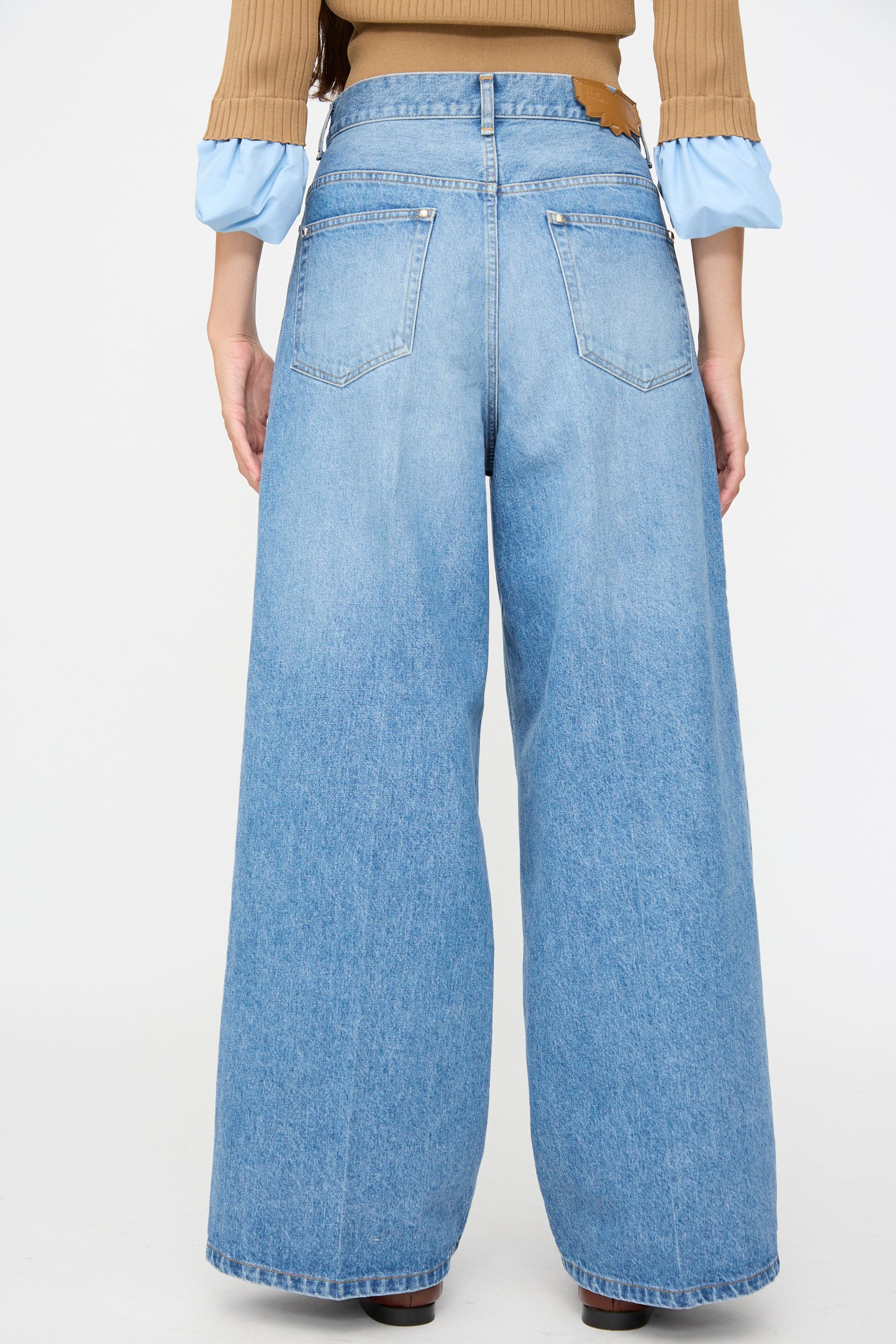 A person is shown from the back against a plain white background, wearing TOGA ARCHIVES Denim Wide Pants in Blue styled with a brown ribbed sweater featuring blue cuffs.