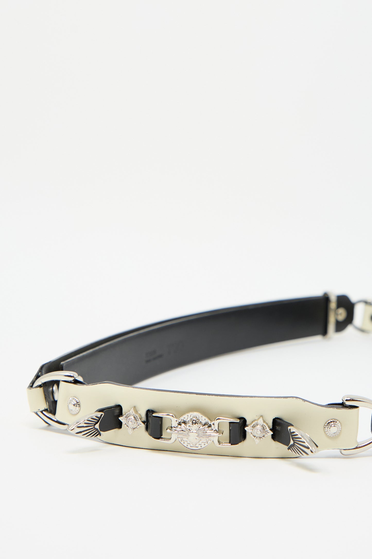 The Metal Leather Narrow Belt in Off White by TOGA ARCHIVES features a silver metal section with diamond-shaped embellishments and wing designs in the center, exuding a Western-inspired charm. This adjustable belt seamlessly combines style and functionality.