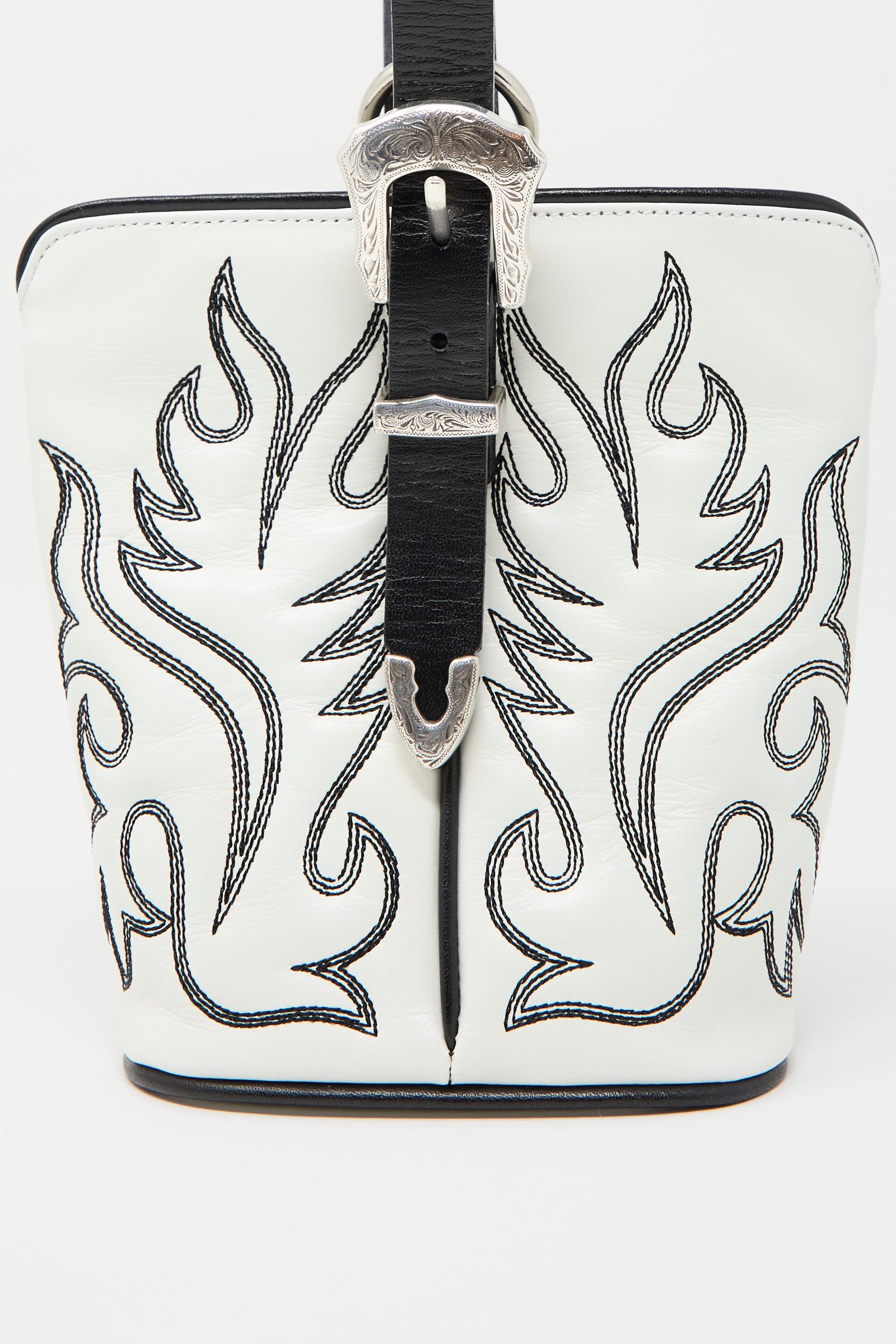 The Western Leather Handbag in White by TOGA ARCHIVES is a small structured leather bag made of white cowhide, adorned with black western motif embroidery and featuring a silver buckle on the strap.