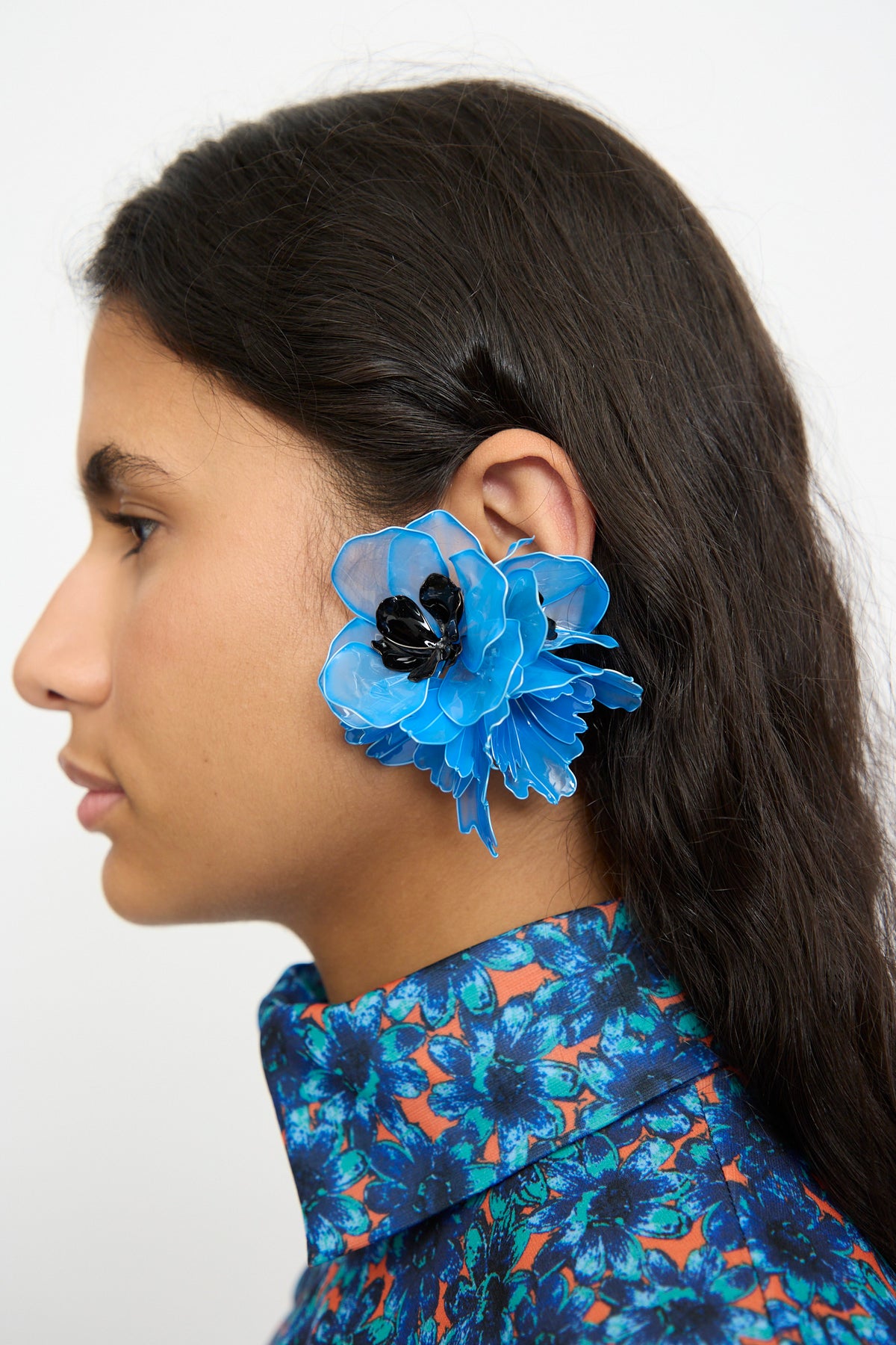 A close-up side profile of a woman with long dark hair wearing TOGA ARCHIVES Flower Earrings in Light Blue, and a blue, orange, and black patterned shirt against a plain white background.