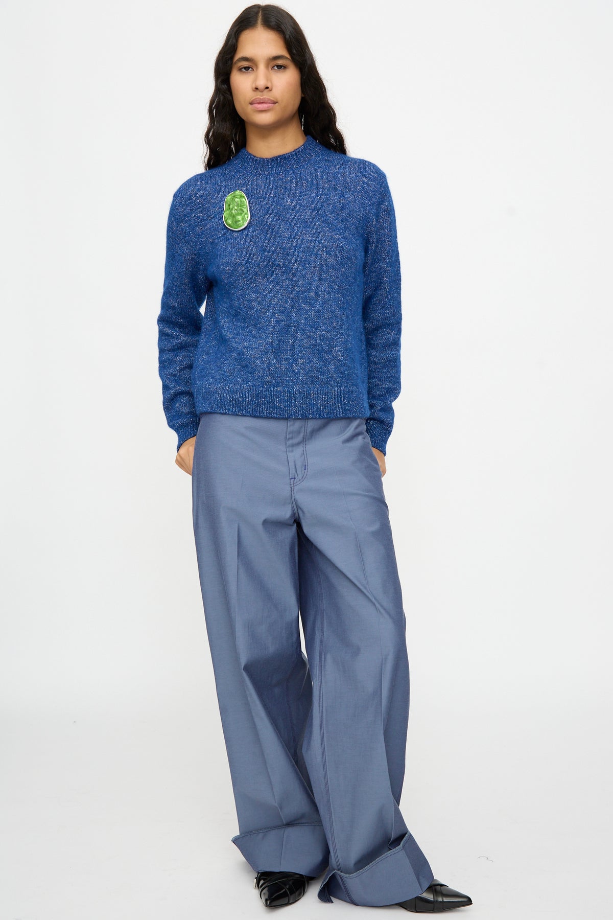 A person wearing a blue sweater styled with TOGA ARCHIVES’ Round Wave Brooch in Light Green and wide-legged light blue pants stands against a plain white background. Crafted in Japan, the silver-tone brass brooch features elegant resin detailing, adding a refined touch to the ensemble—ideal for those who appreciate unique, artisanal accessories.