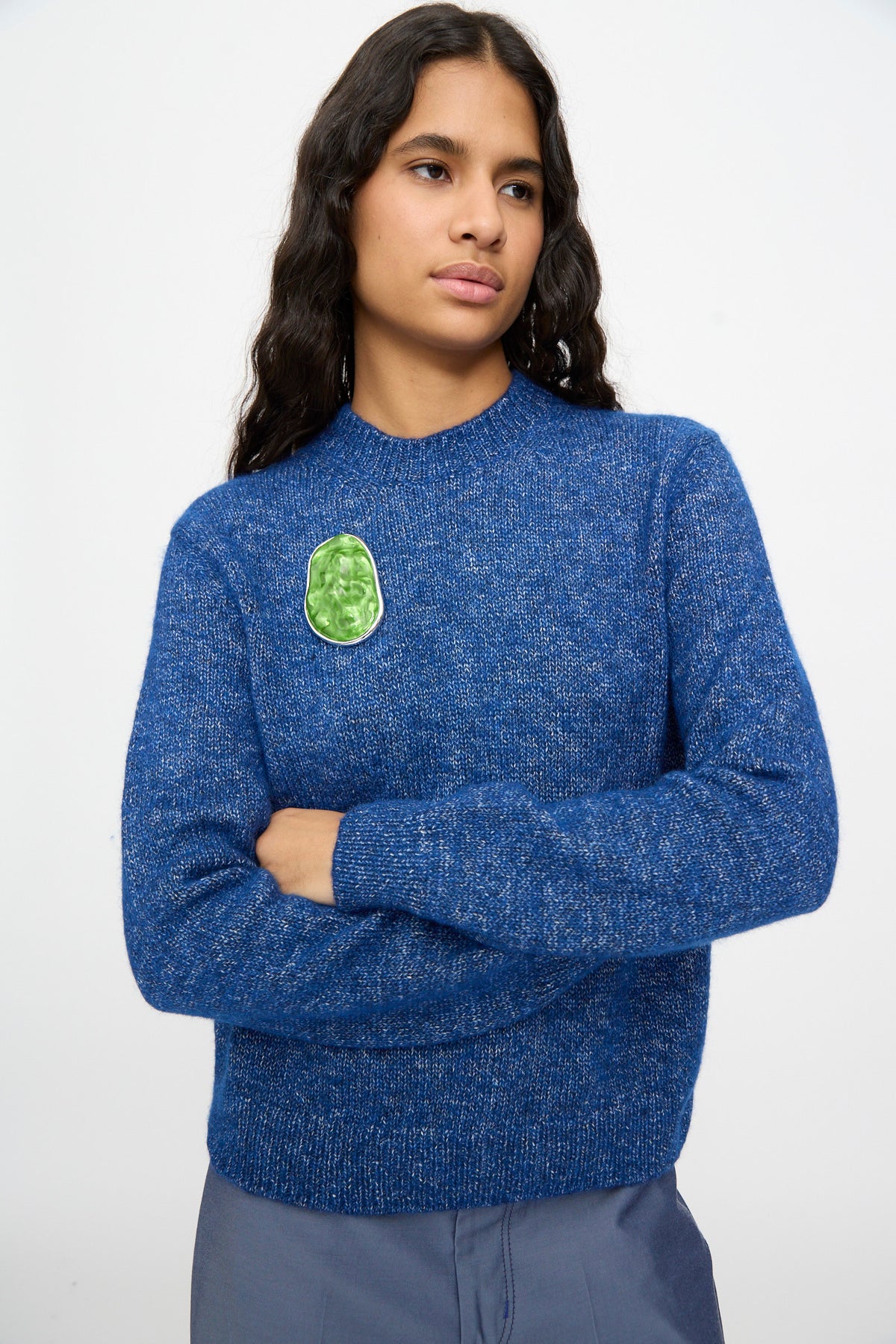 Person with long dark hair wearing a blue sweater with a green patch on the chest, standing with arms crossed against a plain background, accentuated by the TOGA ARCHIVES Round Wave Brooch in Light Green—crafted from silver-tone brass and made in Japan.