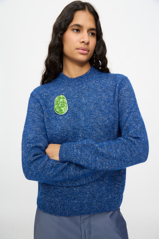 Person with long dark hair wearing a blue sweater with a green patch on the chest, standing with arms crossed against a plain background, accentuated by the TOGA ARCHIVES Round Wave Brooch in Light Green—crafted from silver-tone brass and made in Japan.