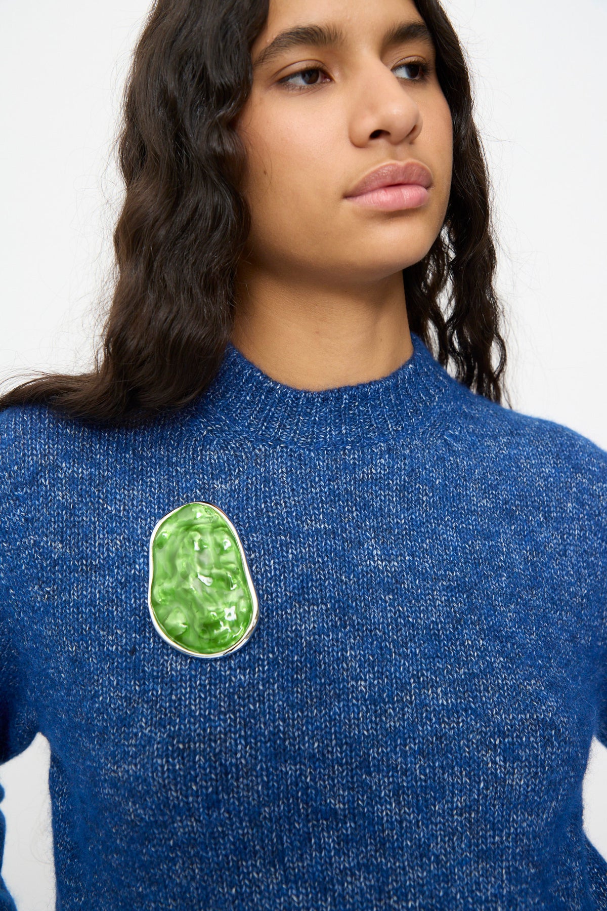 A person wearing a blue knit sweater stands against a plain background with the TOGA ARCHIVES Round Wave Brooch in Light Green—crafted from silver-tone brass and resin in Japan—pinned to their chest.