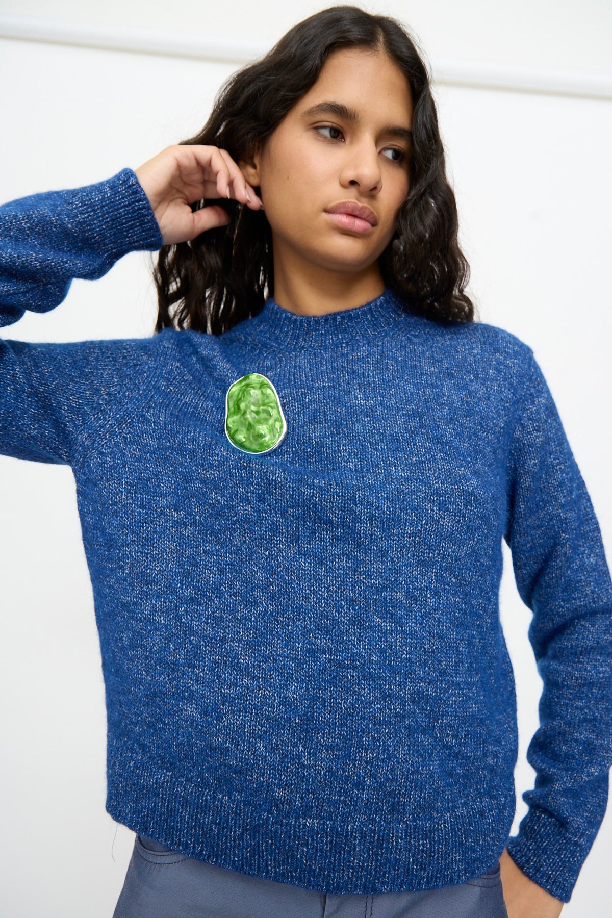 A person wearing a blue knit sweater with a green decorative patch on the left side of the chest, accented by the TOGA ARCHIVES Round Wave Brooch in Light Green, stands against a plain light-colored background.