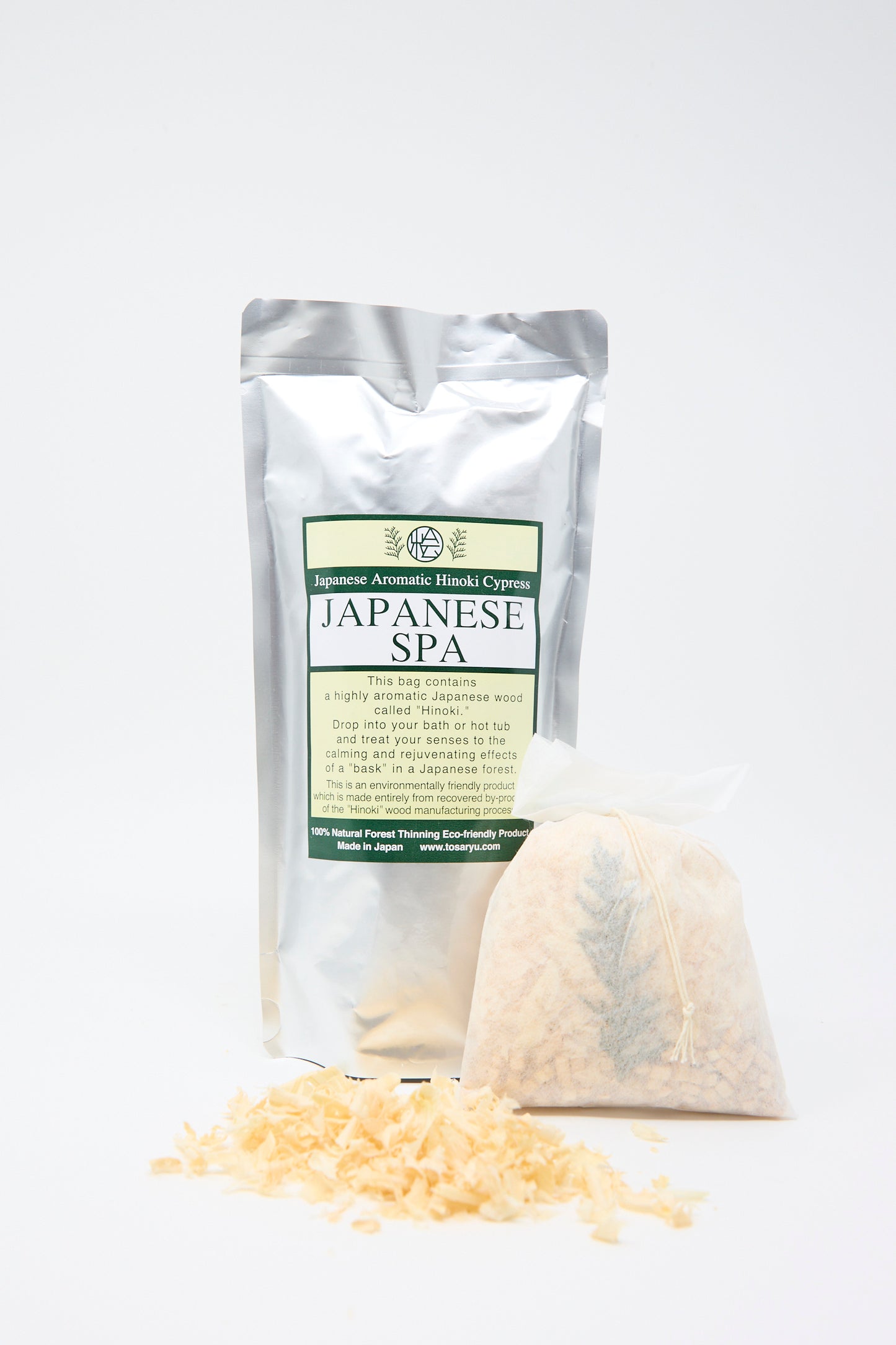 A silver foil package labeled "Hinoki Bath Flakes" by Saikai sits elegantly beside a sachet and aromatic shavings of Hinoki Cypress wood, all against a white background. 