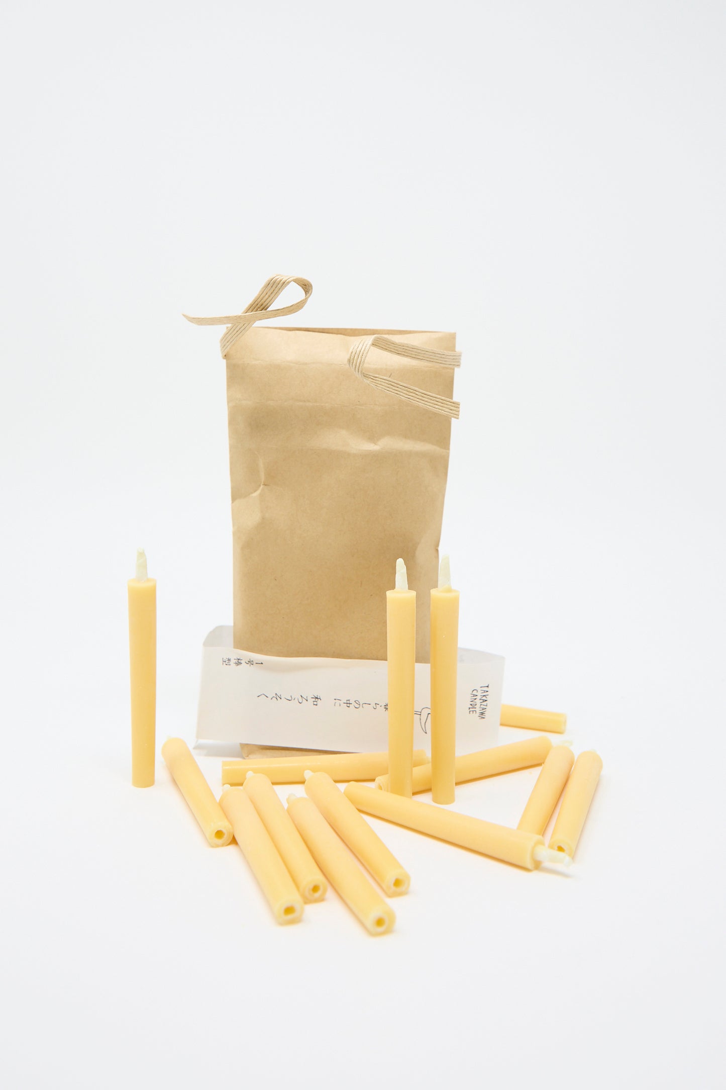 The Komenuka Rice Bran Candle by Saikai is arranged on a white background, featuring three standing upright and others scattered around, all presented in front of a brown paper bag. 