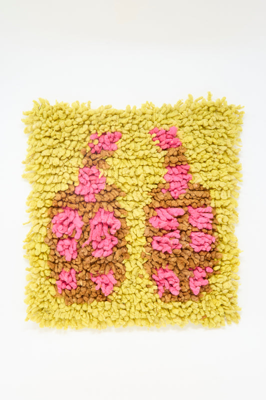 The Chair Rug in Date Fruits by Tamar Mogendorff is a square, handwoven rug with a yellow background and abstract bottle shapes in hot pink and mocha.