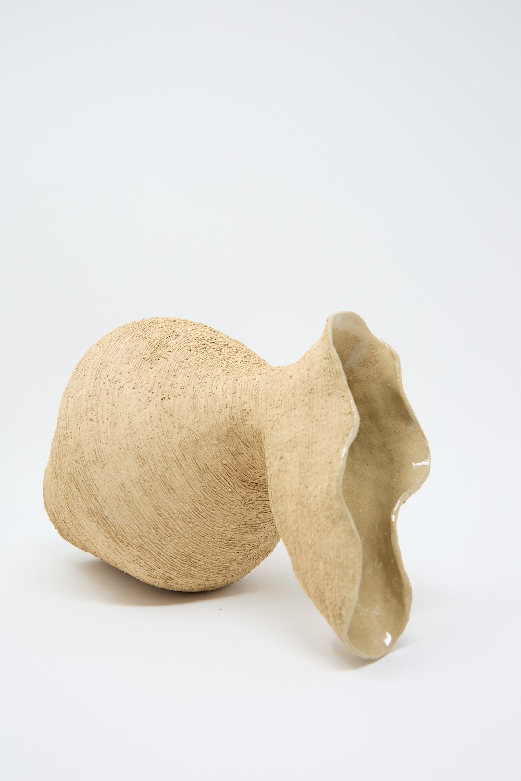 A beige, hand-built vase made from textured sand stoneware lies on its side against a white background, showcasing its wide, wavy-rimmed opening and raw surface finish. The Flutter Vase in Sand by Tania Whalen.