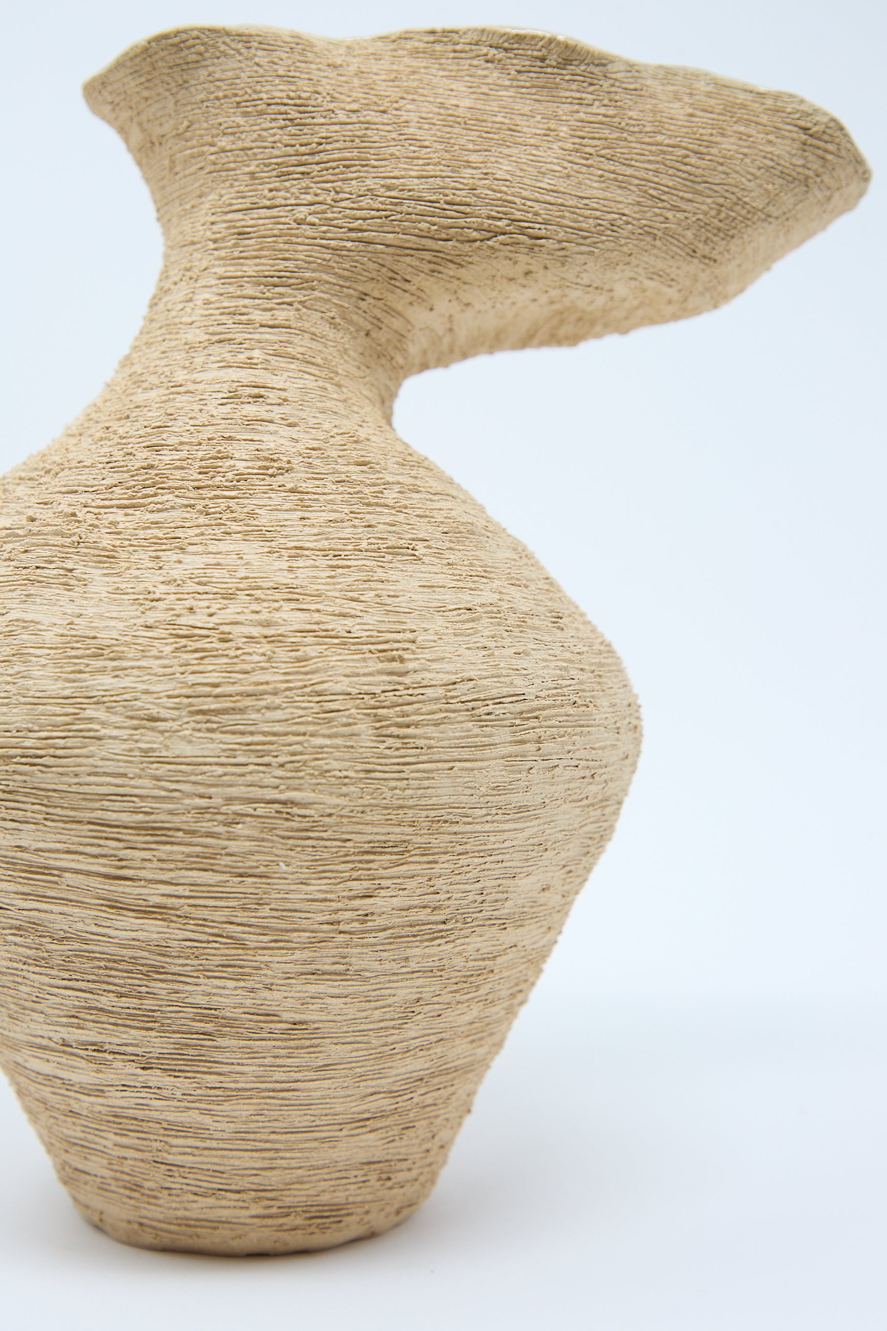 A hand-built vase with a textured, asymmetrical beige design features a unique, curving shape on a plain white background. The raw surface finish highlights the artisanal quality of The Flutter Vase in Sand by Tania Whalen.