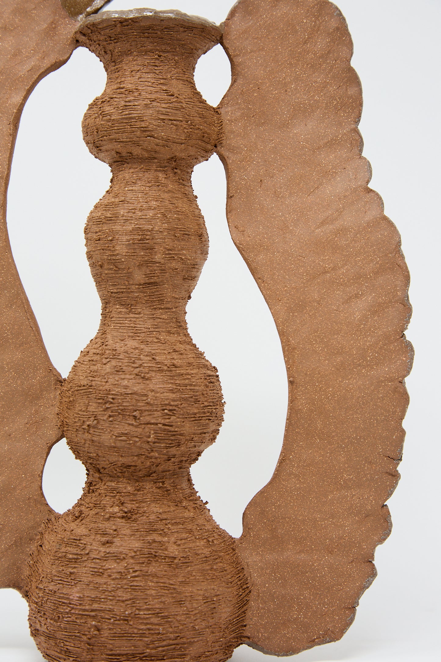 Close-up of a textured, abstract clay sculpture featuring a central column with stacked bulbous shapes and two wing-like extensions on either side, reminiscent of Tania Whalen's The Flying Ra Vase. The predominantly brown sculpture showcases intricate detailing and craftsmanship.