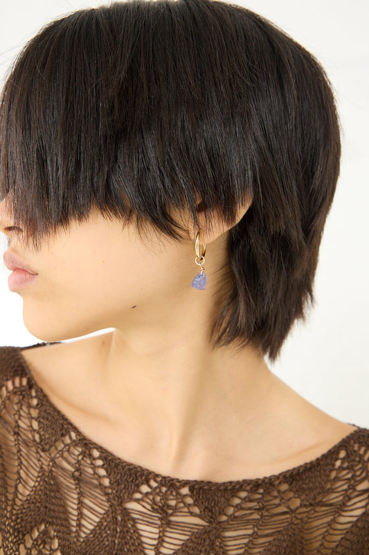 A woman with short hair wearing a brown top and earrings, adorned with a stylish Tara Turner Raw Tanzanite Hoop Earrings pendant.
