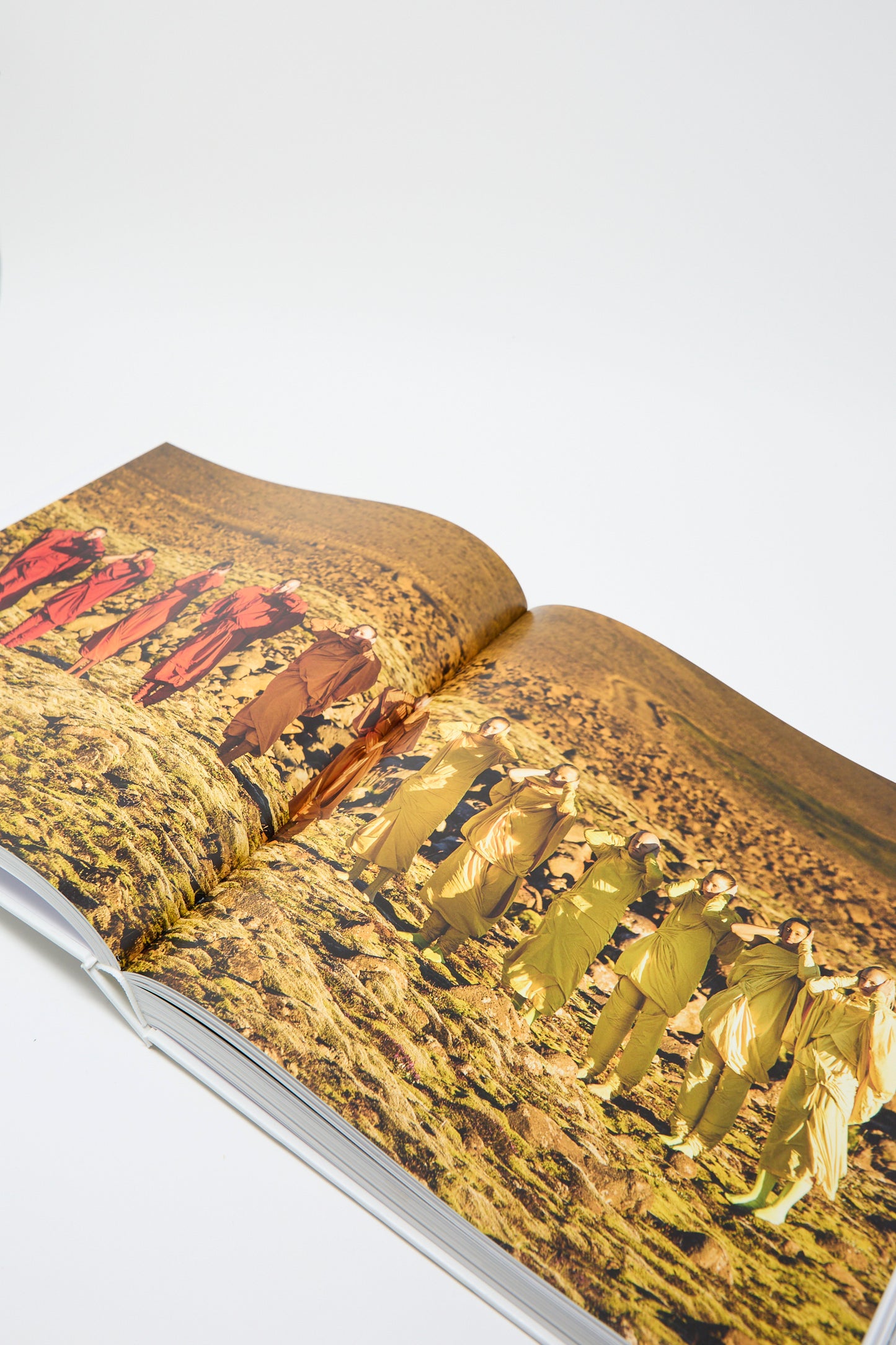 An open page of the book, Issey Miyake by Taschen, reveals a photograph of people dressed in colorful outfits against a barren landscape, placed on a white background. 