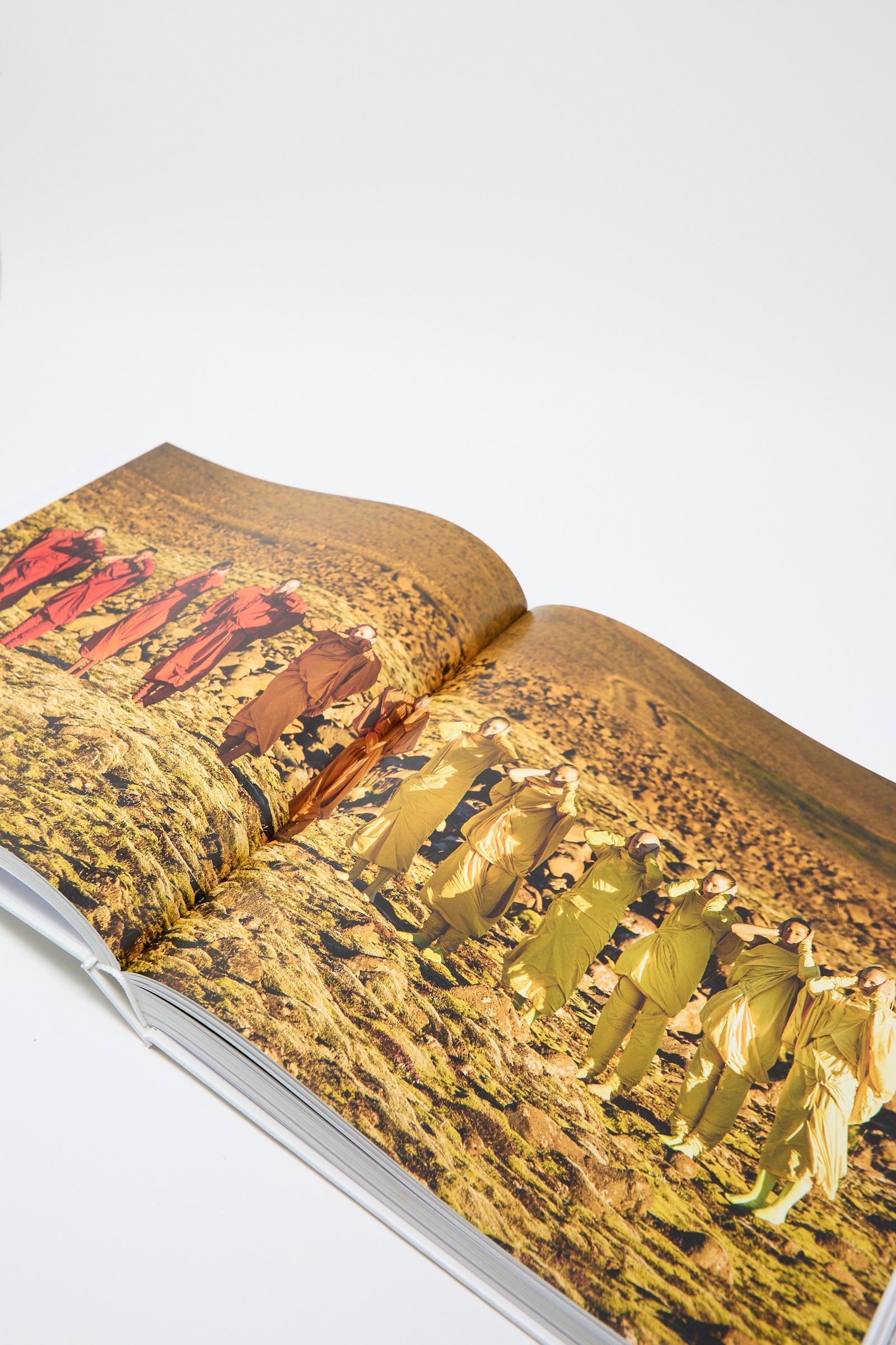 An open page of the book, Issey Miyake by Taschen, reveals a photograph of people dressed in colorful outfits against a barren landscape, placed on a white background. 