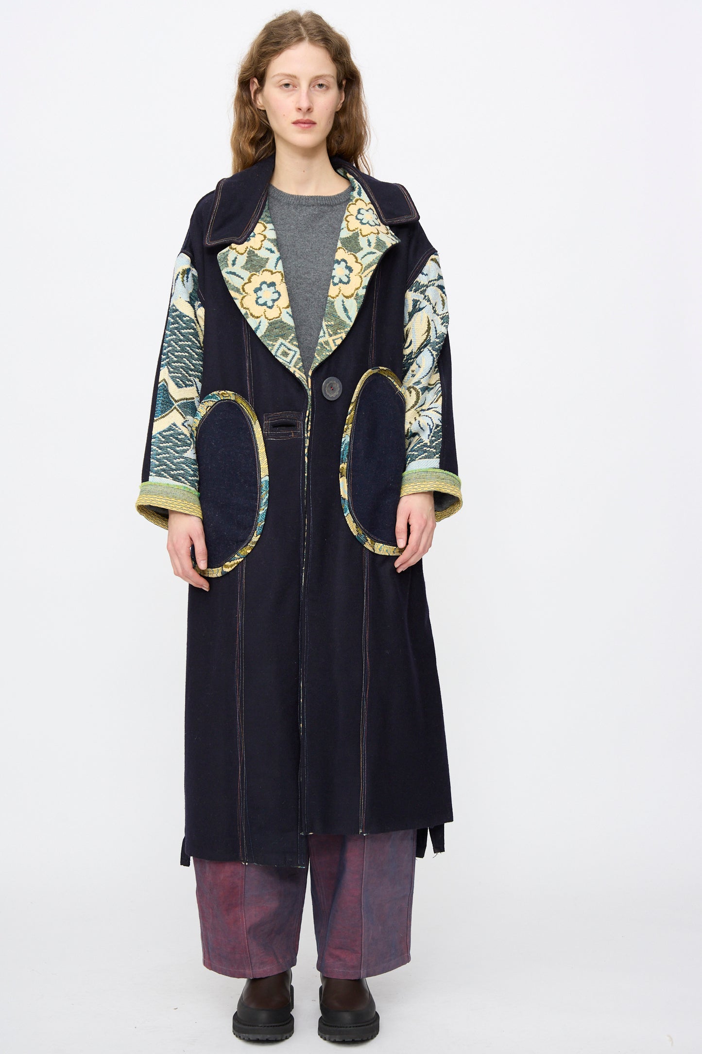 A person stands facing forward in the Thank You Have A Good Day Crosby Coat, an oversized handmade long coat with floral lapels and sleeves, paired with a grey shirt, wide red pants, and black shoes against a plain white background.