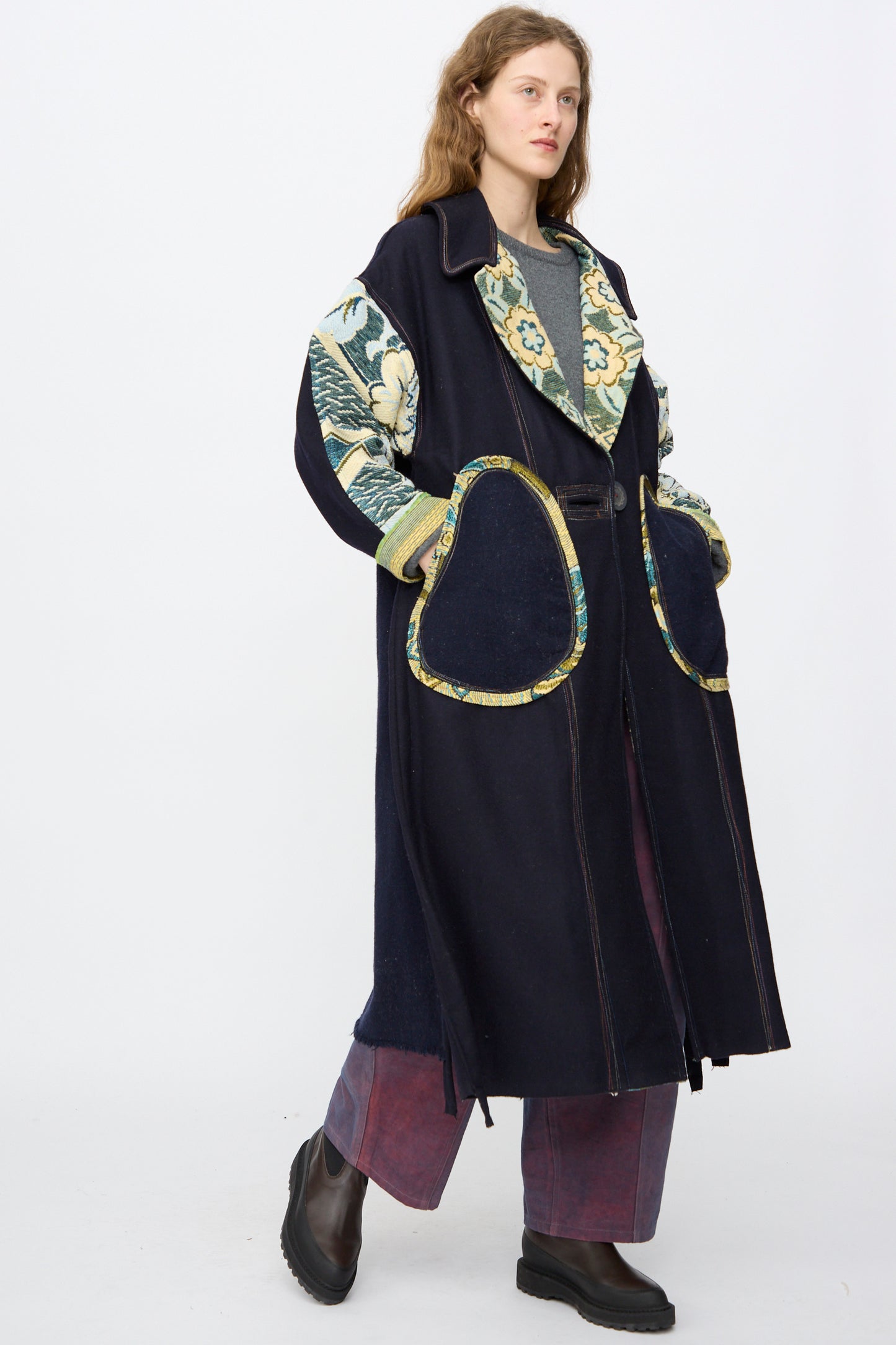 A person models the oversized, handmade Crosby Coat by Thank You Have A Good Day, featuring floral-patterned sleeves and trim, large pockets, wide-leg pants, and black boots against a plain white background.