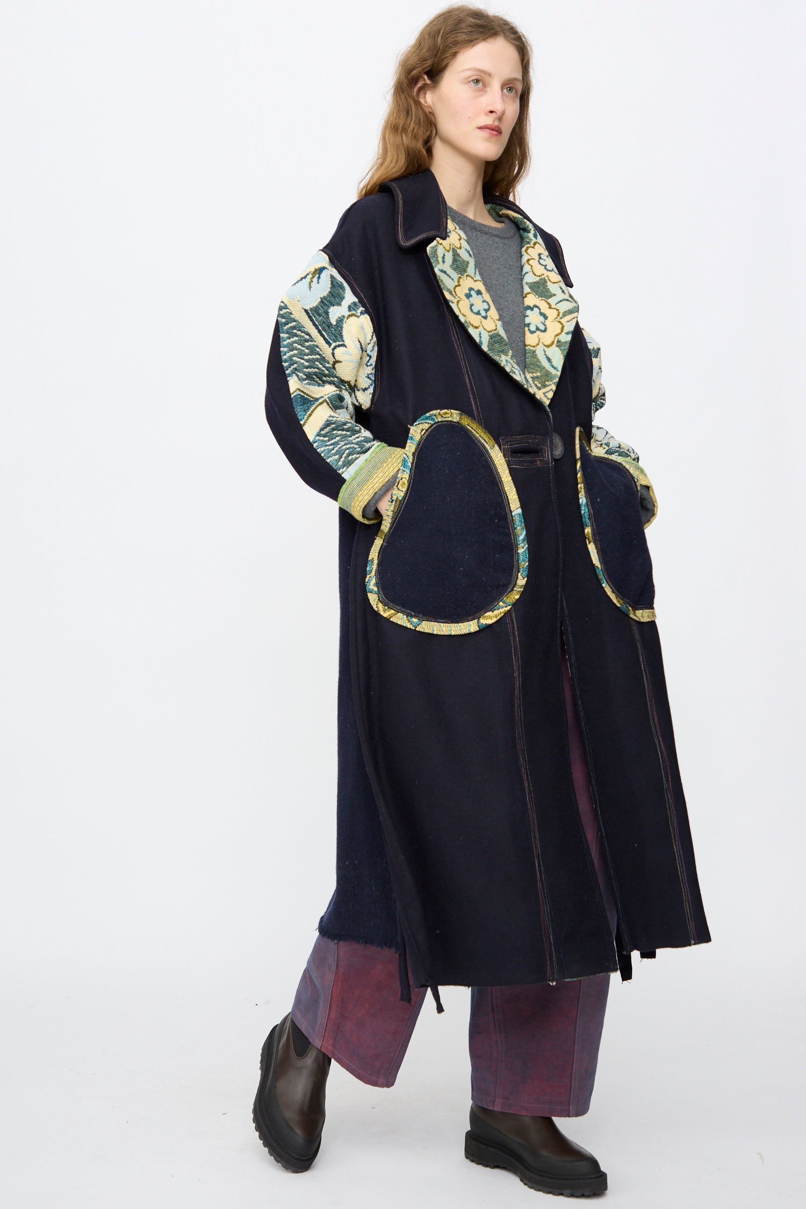 A person models the oversized, handmade Crosby Coat by Thank You Have A Good Day, featuring floral-patterned sleeves and trim, large pockets, wide-leg pants, and black boots against a plain white background.