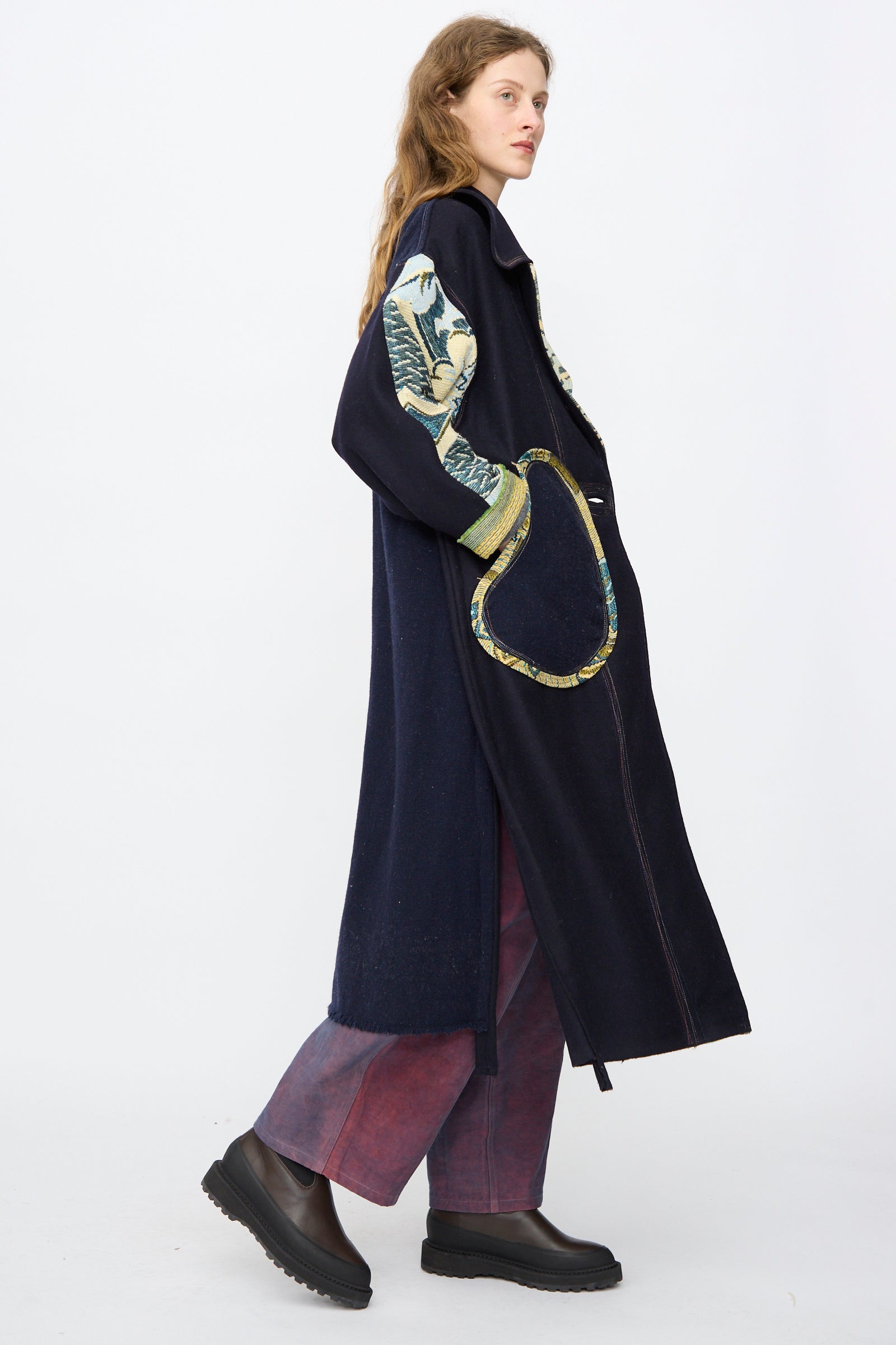 A person stands in profile against a white background, wearing the navy handmade Crosby Coat by Thank You Have A Good Day, featuring patterned sleeves and pocket, paired with wide maroon pants and black platform shoes.