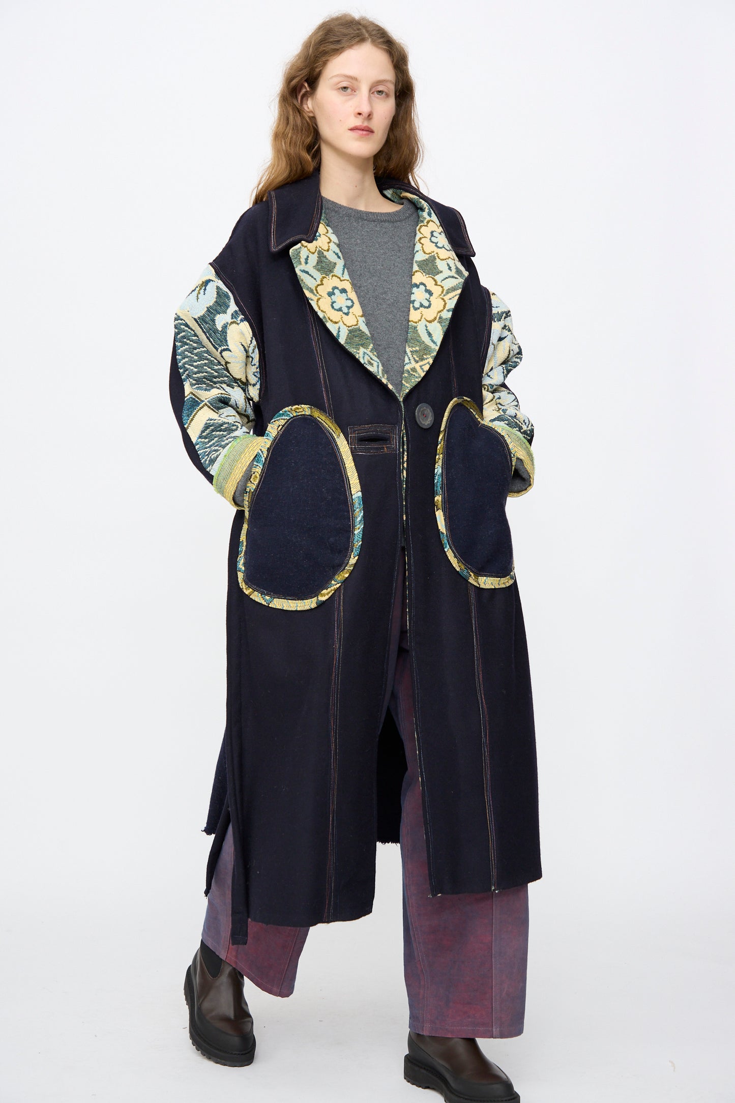 A person stands against a plain background wearing the Crosby Coat by Thank You Have A Good Day, a handmade 100% wool long coat with oversized fit, large floral-patterned sleeves and pockets, paired with a grey top, maroon pants, and dark boots.