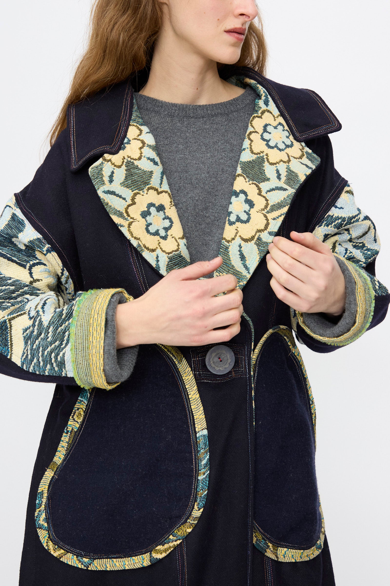 A woman wears the Crosby Coat by Thank You Have A Good Day—an oversized 100% wool coat with patchwork florals, large front pockets, and decorative cuffs—layered over a grey sweater.