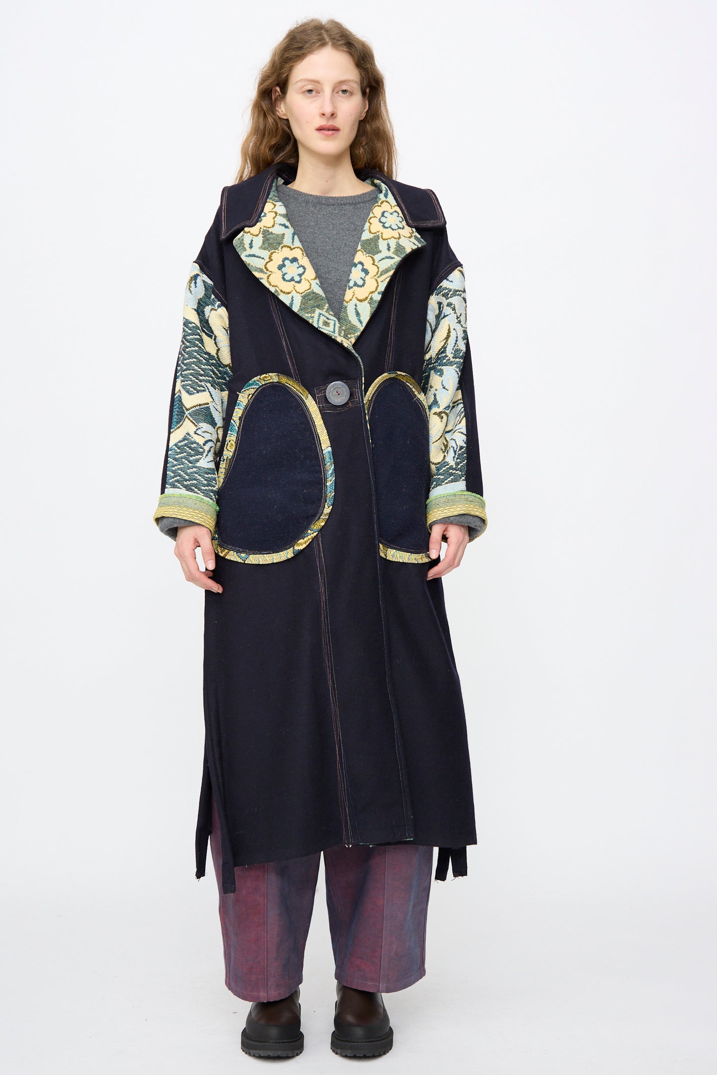 A woman faces forward against a plain background, wearing the oversized Crosby Coat by Thank You Have A Good Day with large decorative pockets and patterned sleeves, paired with a gray top, loose purple pants, and black shoes.