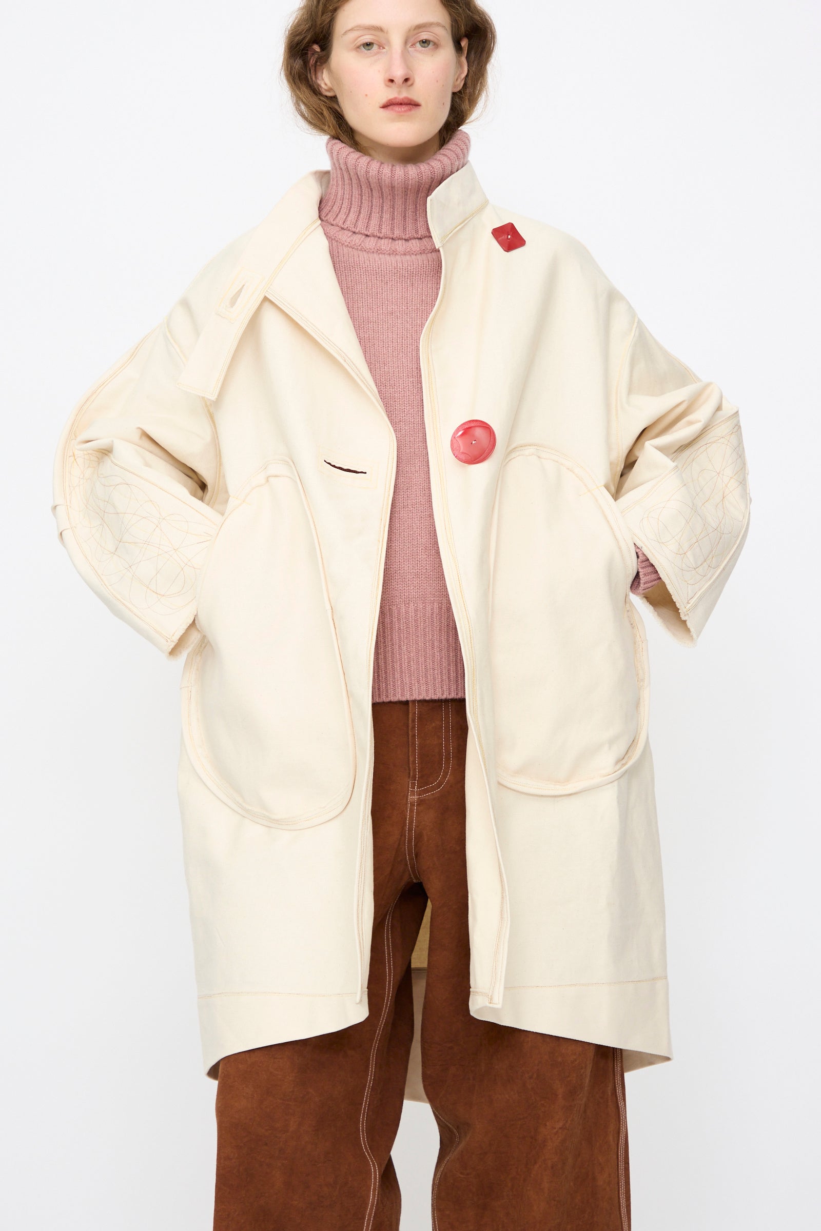 A person wears the Thank You Have A Good Day Denim Fishtail Parka in Ecru, paired with a pink turtleneck sweater and brown pants, standing against a plain white background.
