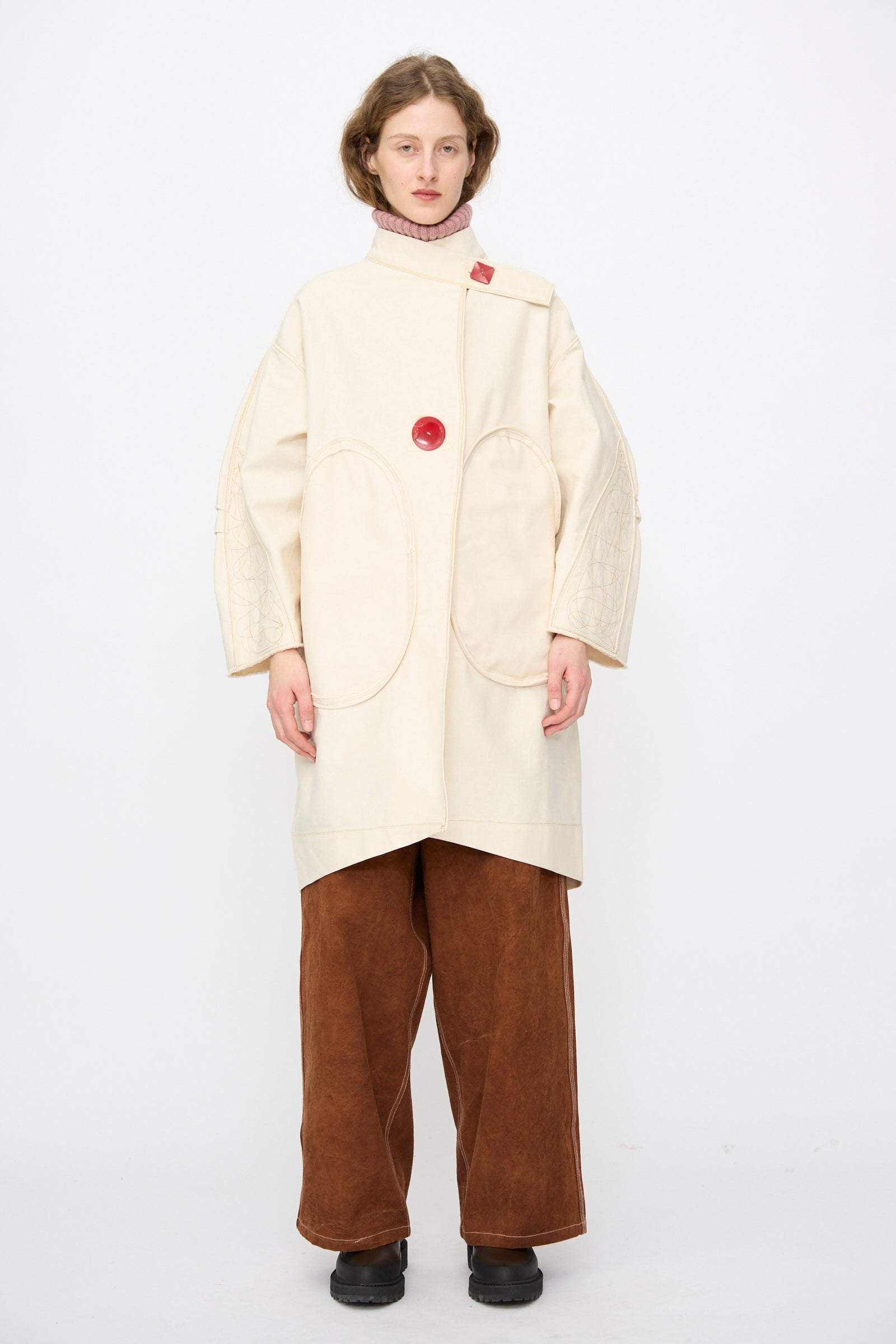 A person stands against a white background wearing the Denim Fishtail Parka in Ecru by Thank You Have A Good Day, featuring an oversized fit, large oval pockets, high collar, red button detail, wide brown pants, and black shoes.