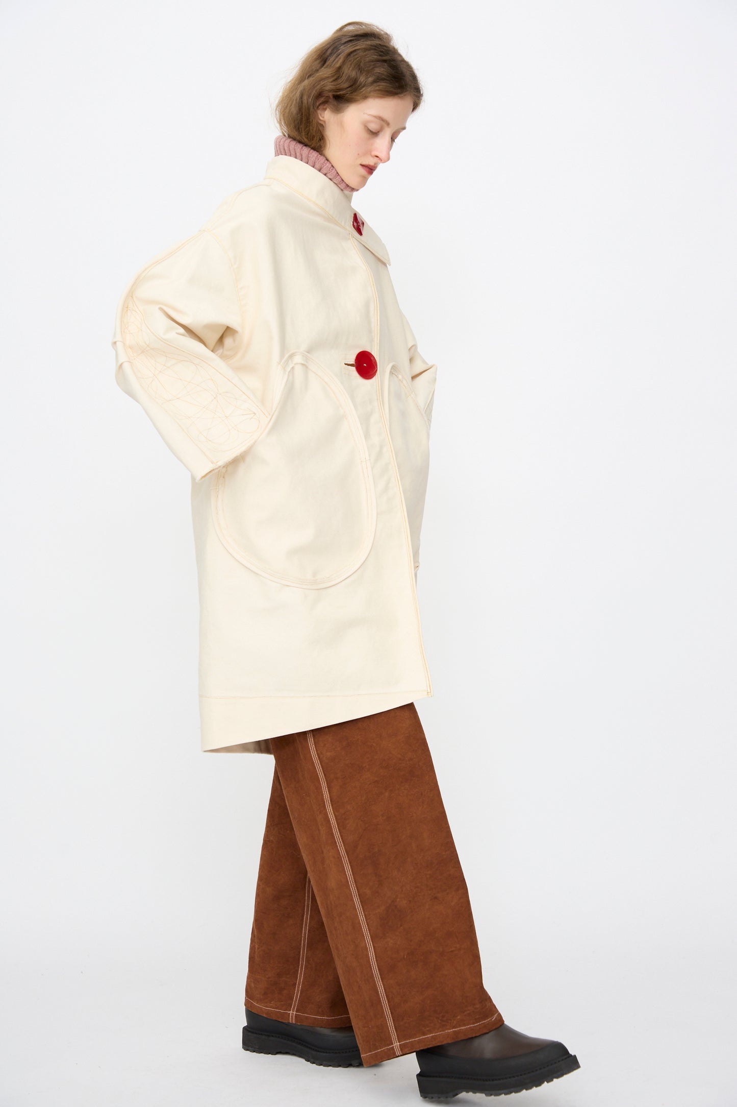 A person stands sideways wearing the Denim Fishtail Parka in Ecru by Thank You Have A Good Day, styled with brown wide-leg pants, black boots, and a pink turtleneck sweater.